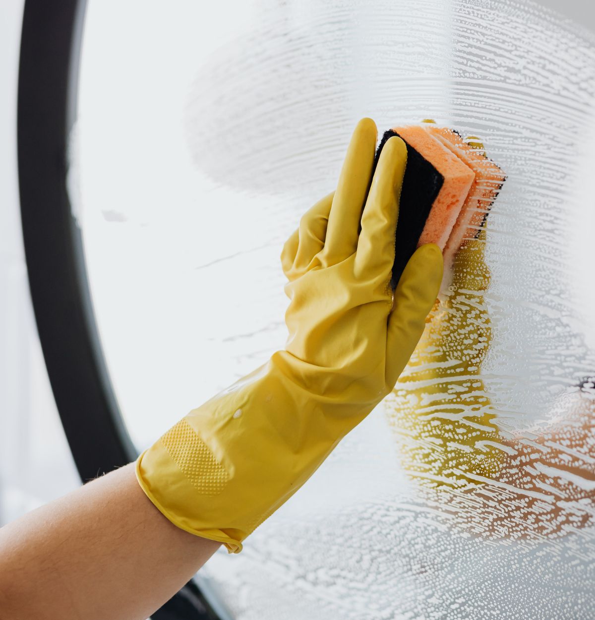 Hand wearing a yellow rubber glove cleaning a glass surface with an orange and black sponge covered in soap suds.