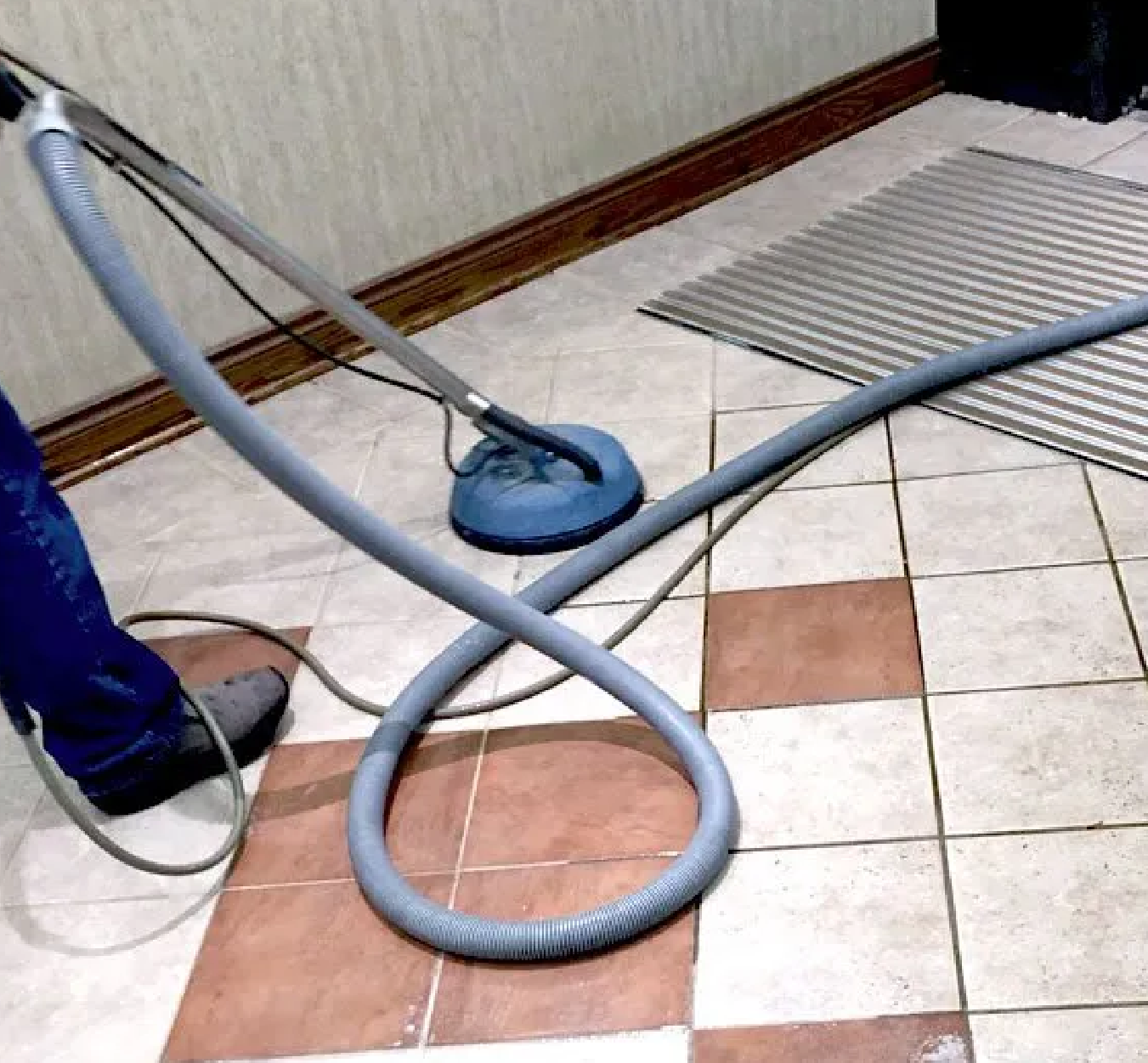 Person using a blue industrial floor cleaning machine on a tiled floor with alternating white and brown square tiles.