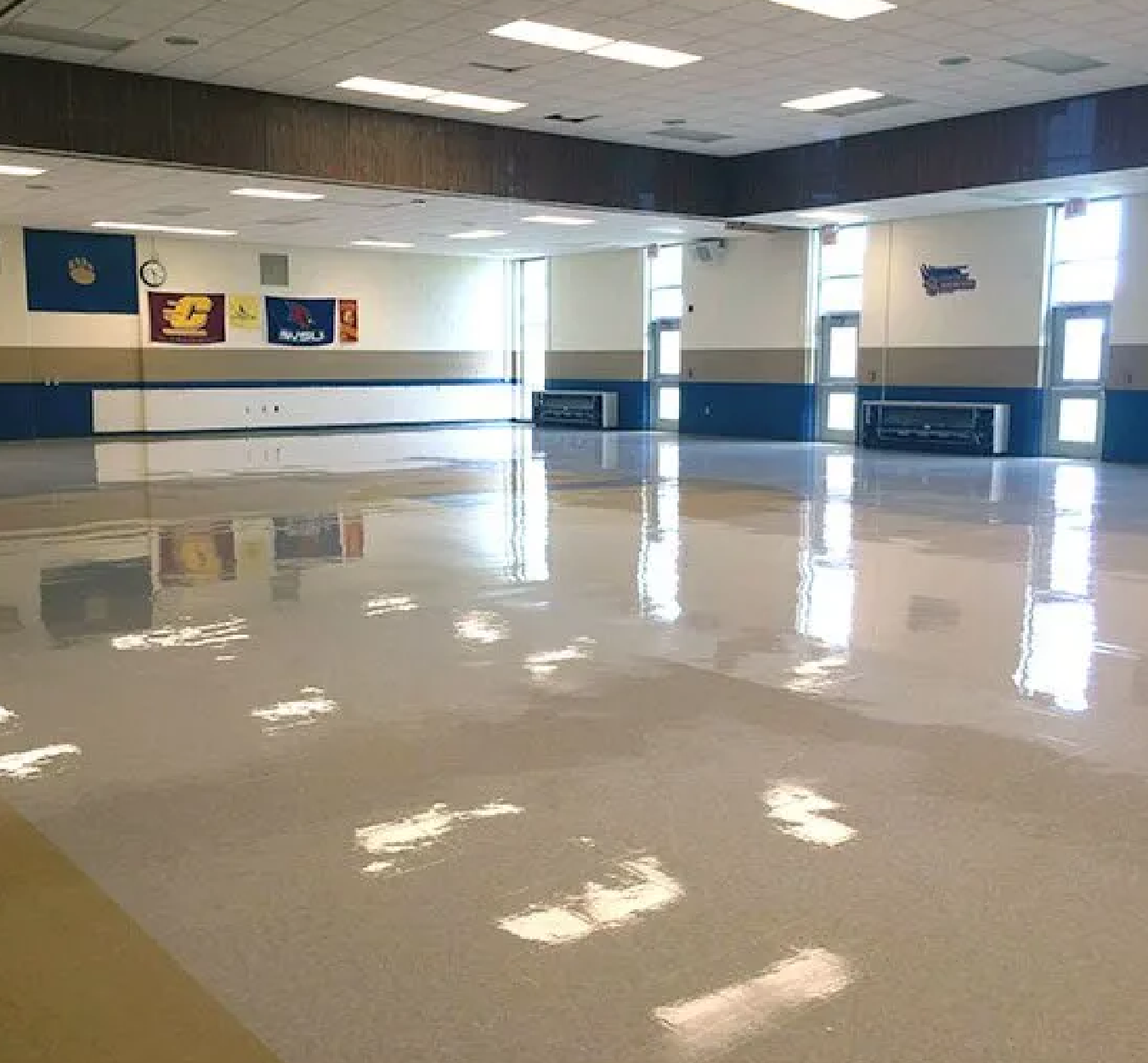 Large empty room with a highly polished floor reflecting ceiling lights, beige and blue walls, and banners with logos on the far wall beneath a clock.
