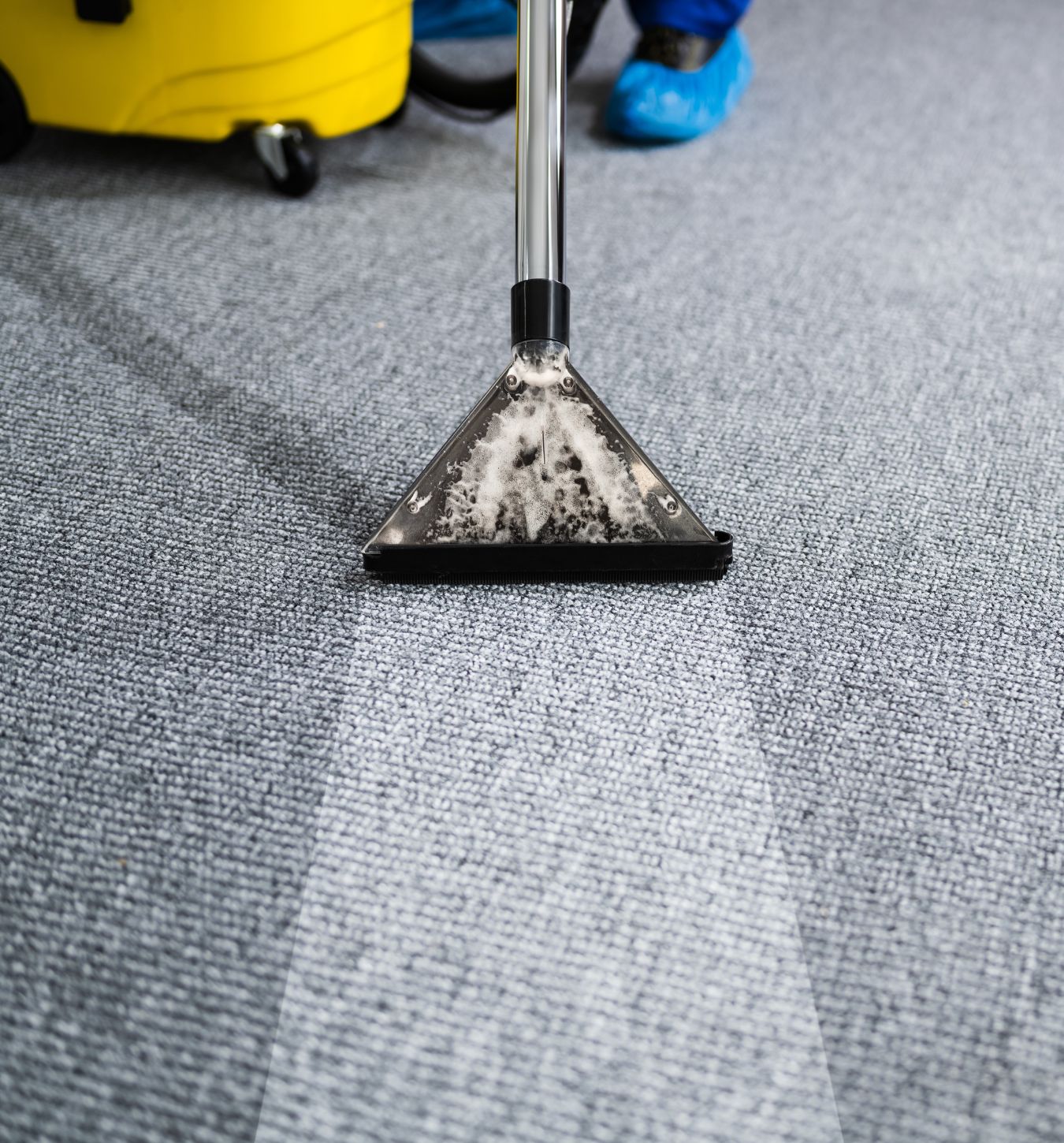 Close-up of a carpet cleaning machine with a transparent triangular head applying foam and cleaning a gray carpet, showing a clean strip behind it.