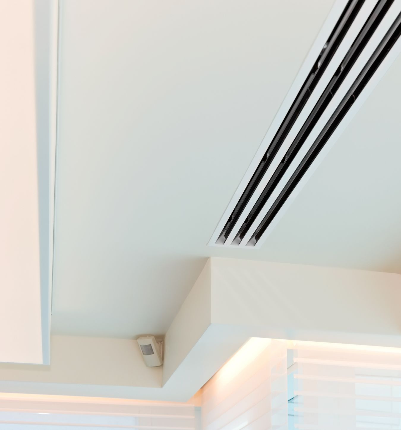 Indoor ceiling corner featuring a white air vent with three narrow black slats, a motion sensor mounted on the wall below the vent, and soft ambient lighting from hidden light strips above translucent window blinds.