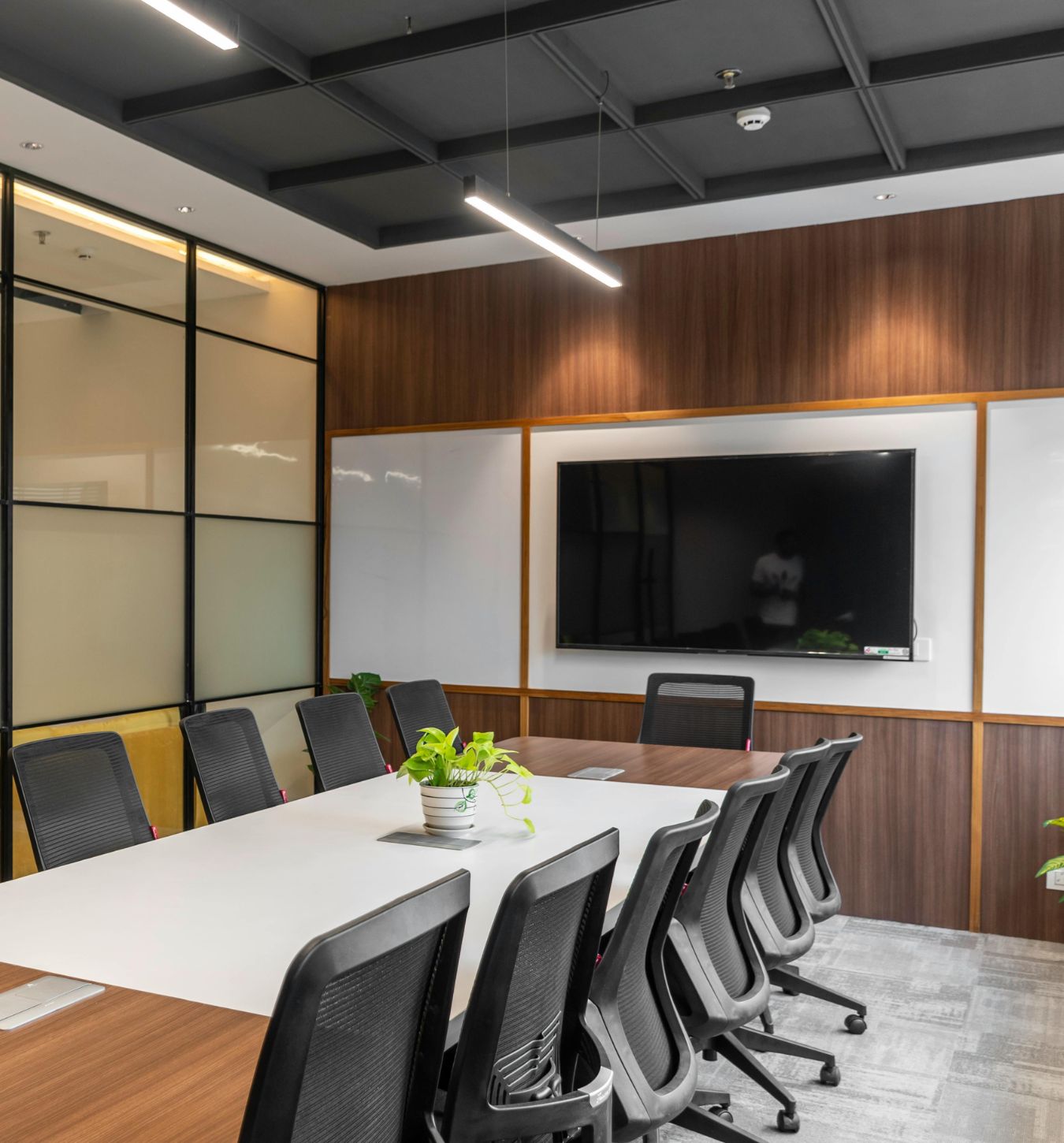Modern conference room with a long white and wood table surrounded by black mesh office chairs, a large flat-screen TV mounted on a wood-paneled wall with whiteboards on either side, and a small green potted plant on the table.