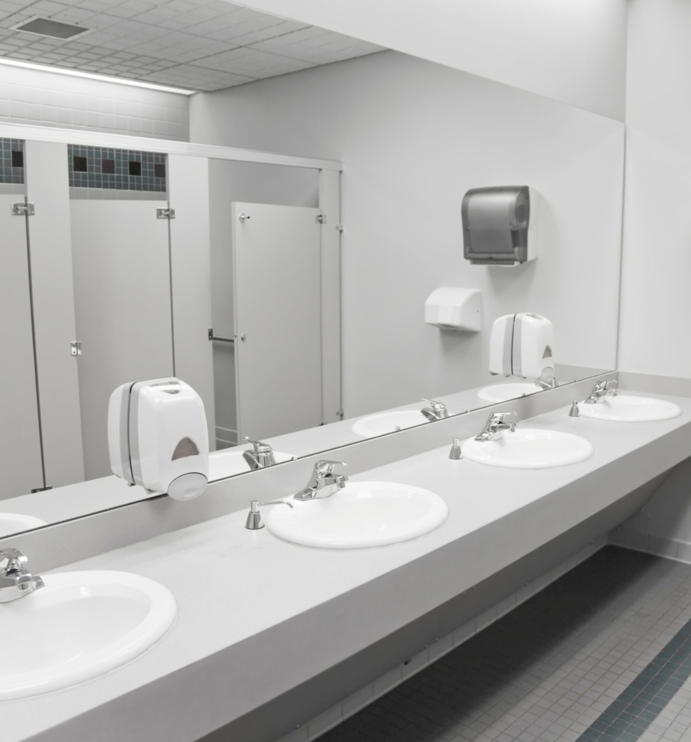 Bright public restroom with a long gray countertop holding four white sinks and silver faucets, large mirror above, white soap dispensers attached to the mirror, and closed white stall doors in the background.