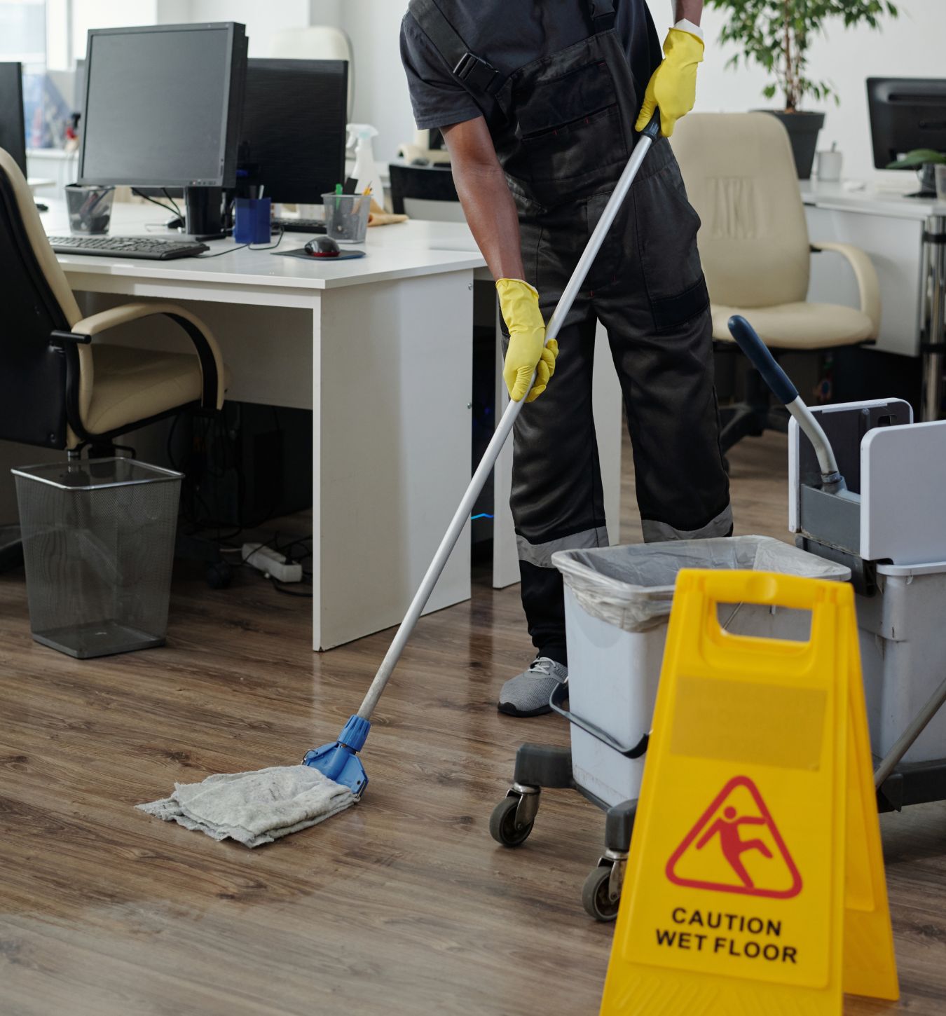 Person wearing yellow gloves and black coveralls mops a wooden office floor near desks and chairs with a yellow caution wet floor sign in the foreground.
