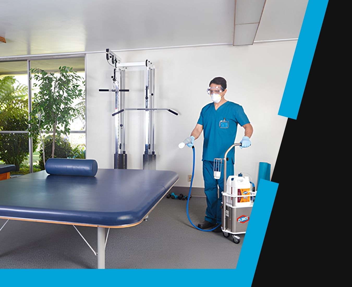 A person in blue medical scrubs, gloves, goggles, and a mask disinfects a treatment table in a clean, bright therapy room with exercise equipment, using a spraying device connected to a cart with cleaning supplies.