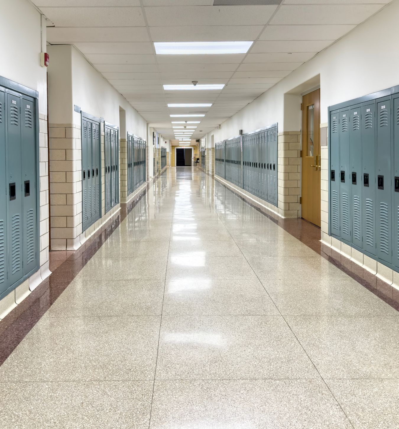 Long empty school hallway with rows of blue lockers on both sides, beige tiled walls, a light brown door on the right, and a reflective tiled floor under ceiling lights.