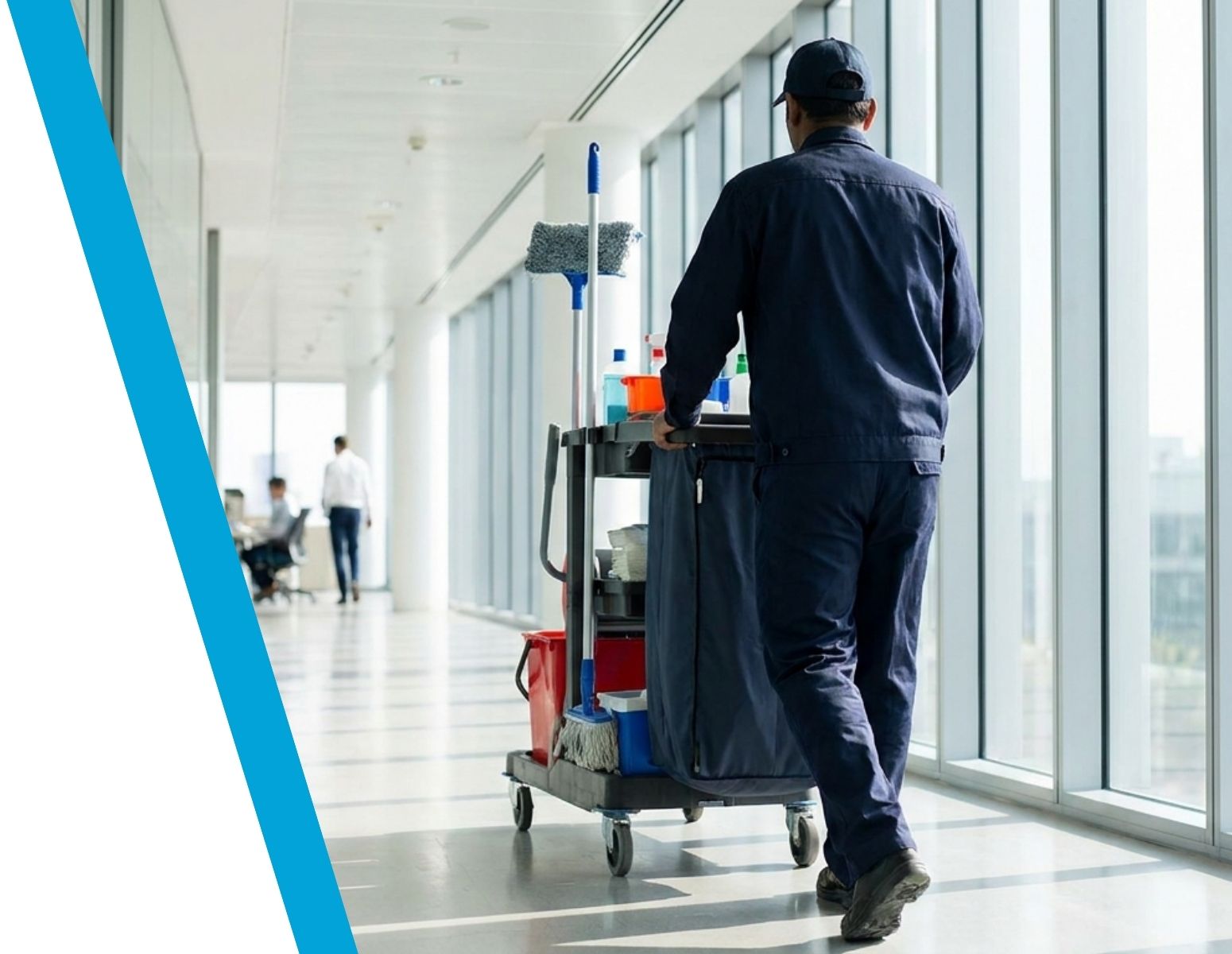 A janitor in a dark uniform pushing a cleaning cart with supplies, mop, and bucket down a bright office hallway with large windows.