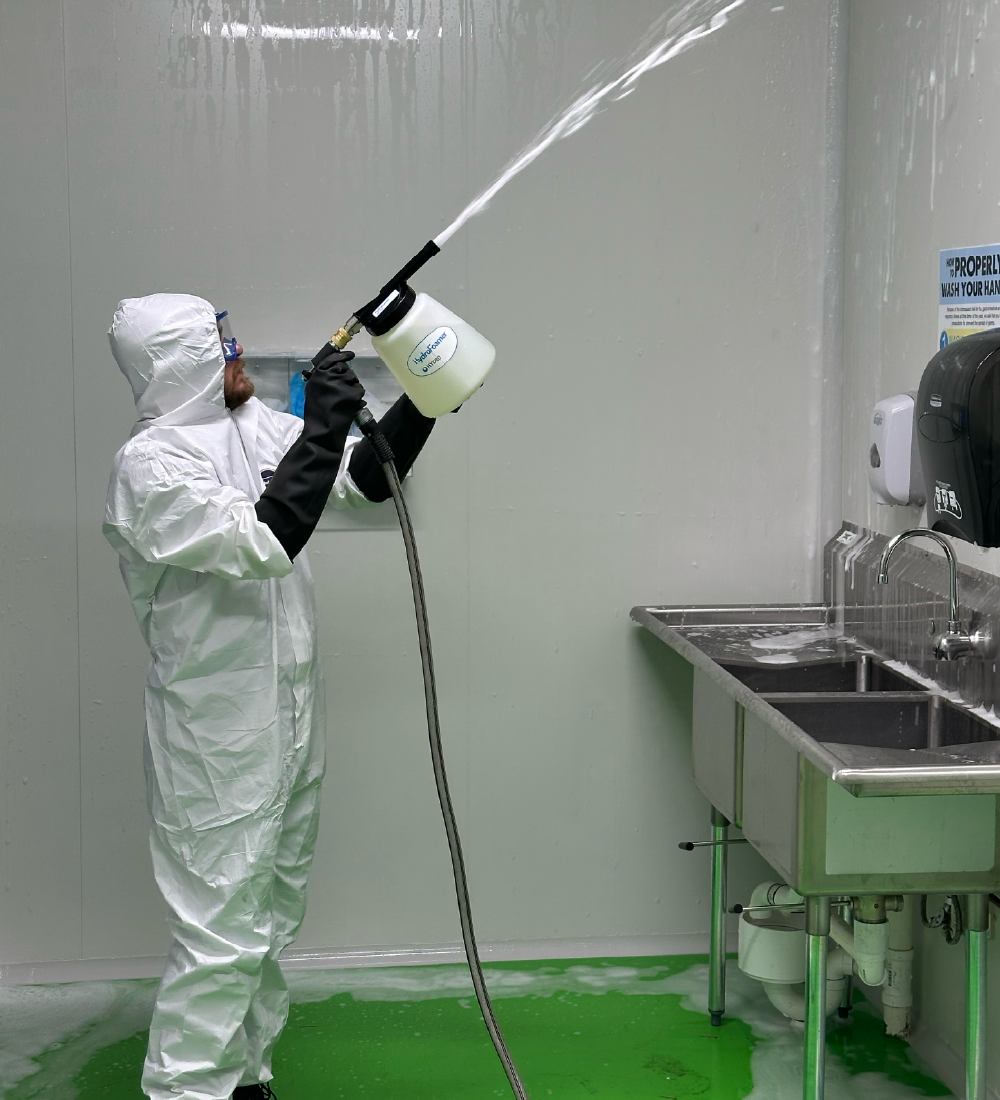 Person in white protective suit, black gloves, and goggles spraying foam sanitizer on a white wall and stainless steel sinks in a sanitation area with a green floor.