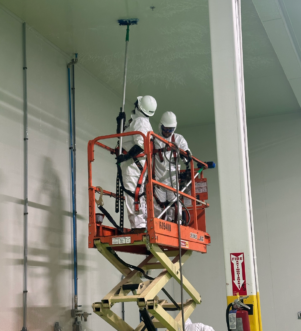Two workers in white protective suits and helmets using an elevated scissor lift inside an industrial building to clean or sanitize the ceiling with brushes on long poles.