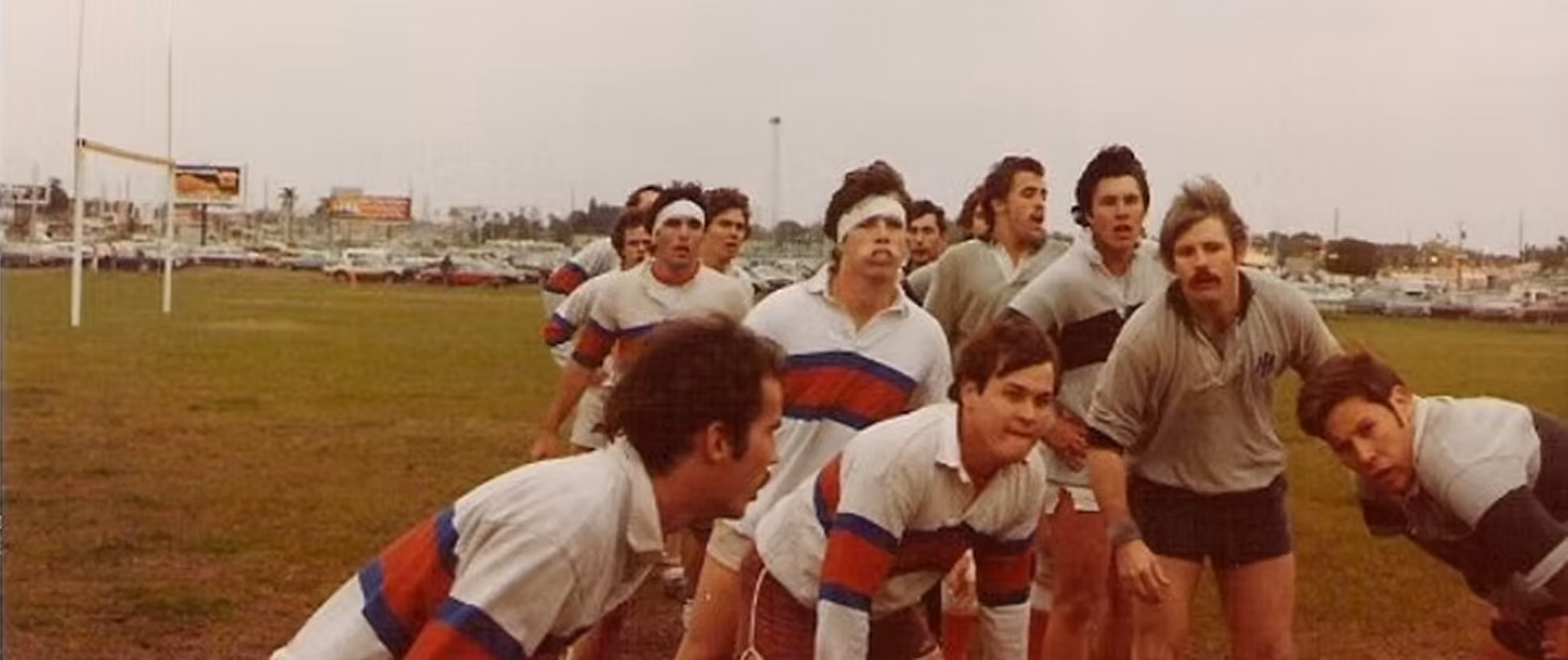 Men in vintage rugby uniforms lined up on a field under overcast sky with rugby goal posts behind.