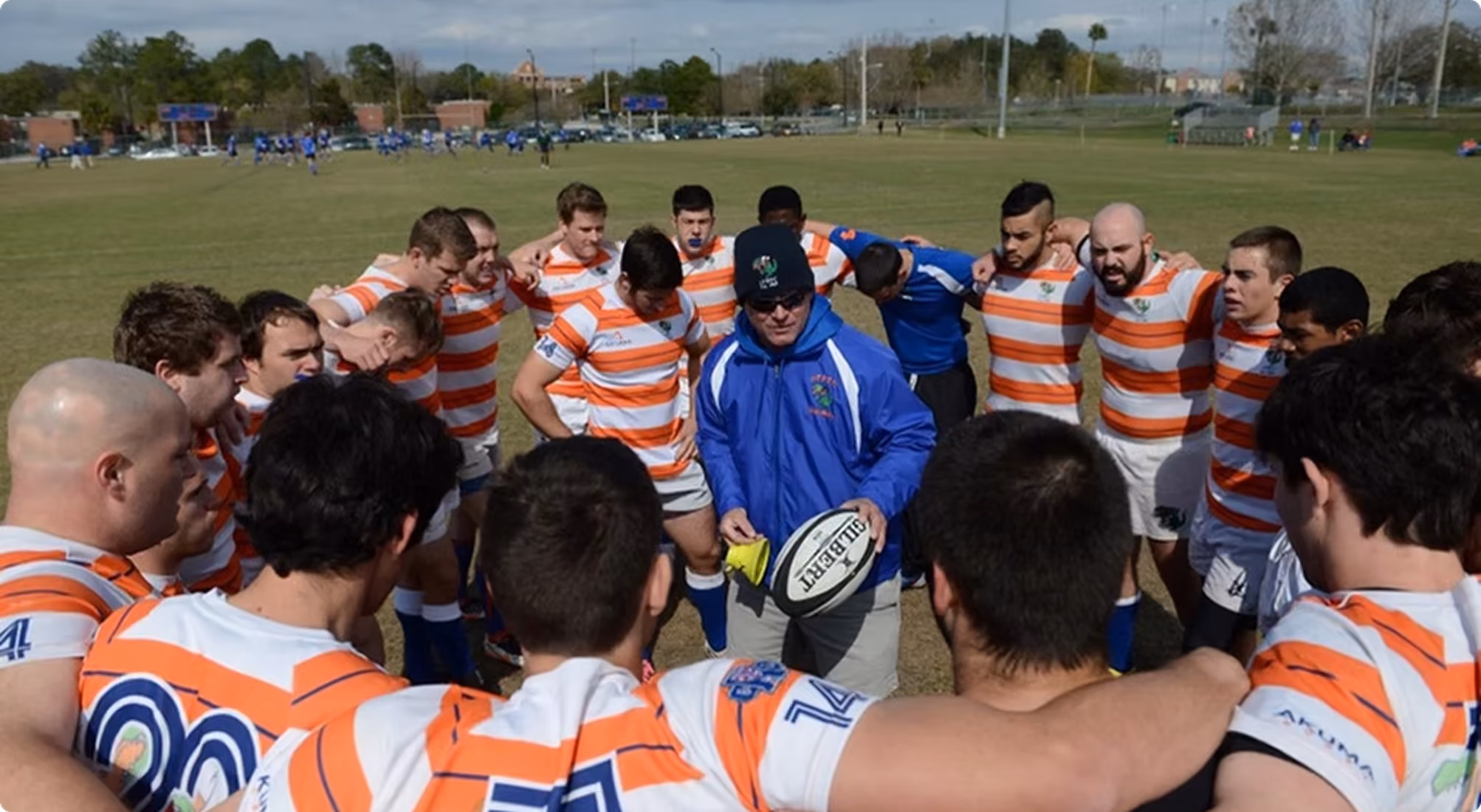 Rugby team dressed in orange and white striped jerseys huddled around coach holding a rugby ball.
