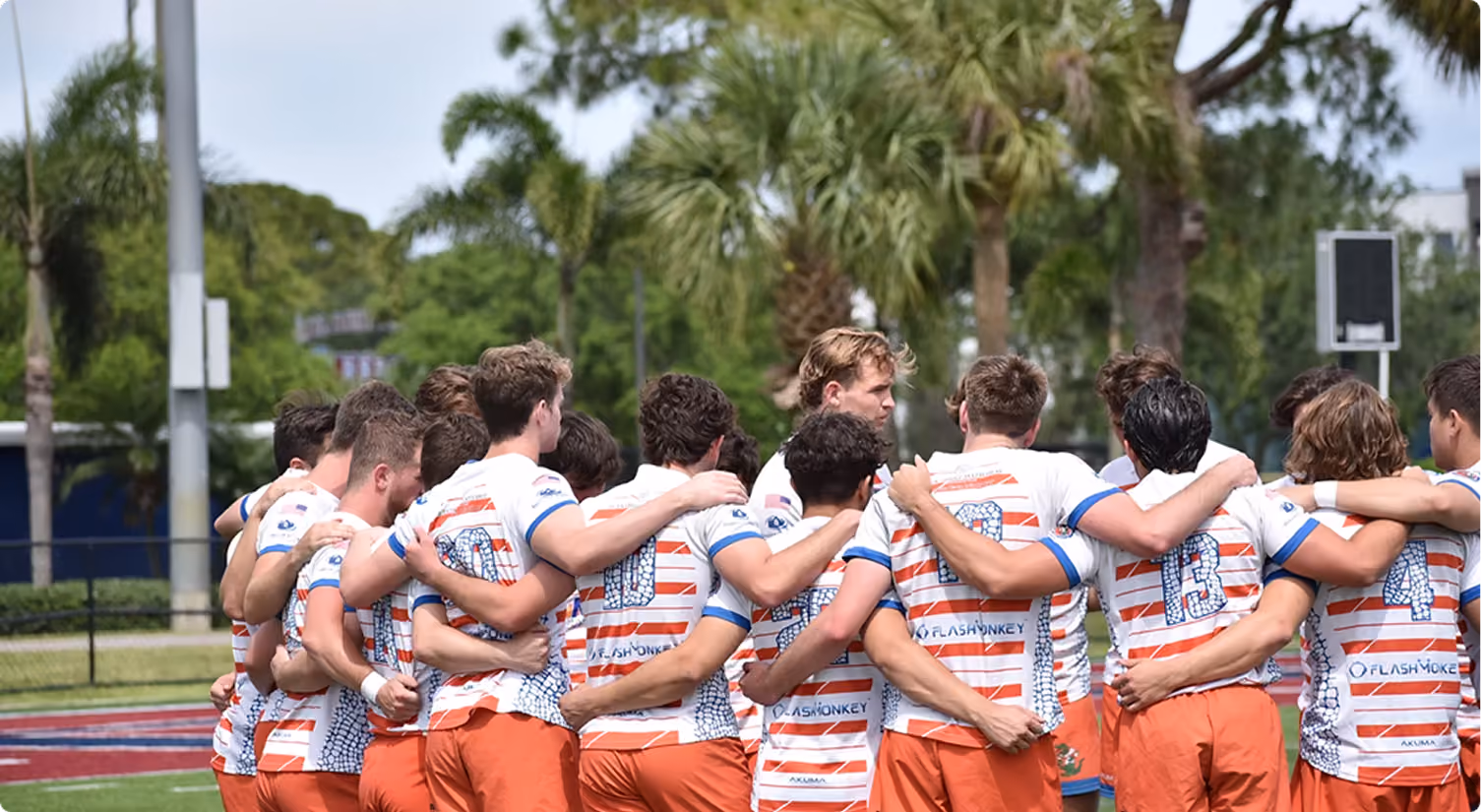 Group of rugby players in striped jerseys and orange shorts huddling on a field with palm trees behind.