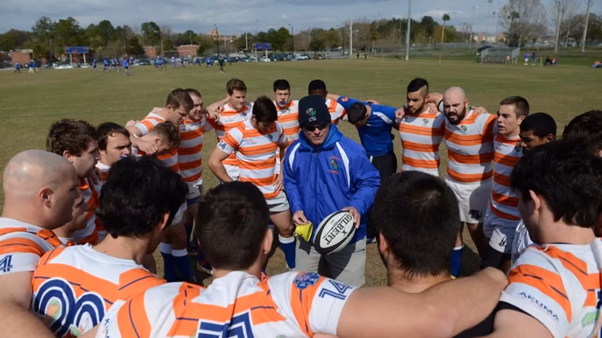 Rugby team in orange-striped jerseys huddled around coach holding a rugby ball on the field.