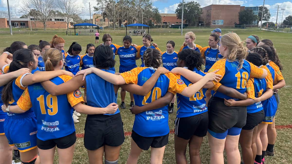 Girls rugby team in blue and orange jerseys huddling with arms around each other on grassy field.