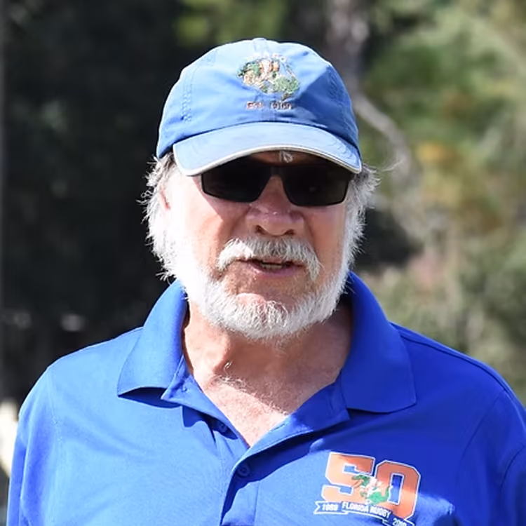 Older man with white beard wearing sunglasses, a blue cap, and a blue shirt outdoors.