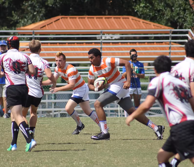 Rugby players in orange-striped and white shirts engaging in play on a grassy field with bleachers in the background.