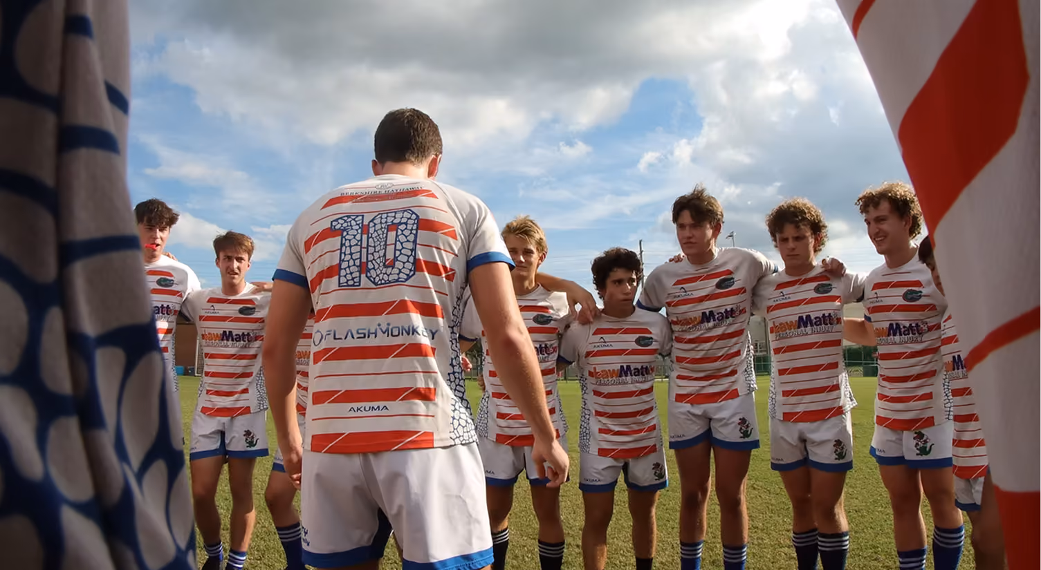 Young rugby players in striped jerseys standing in a huddle on a grass field under a cloudy sky.