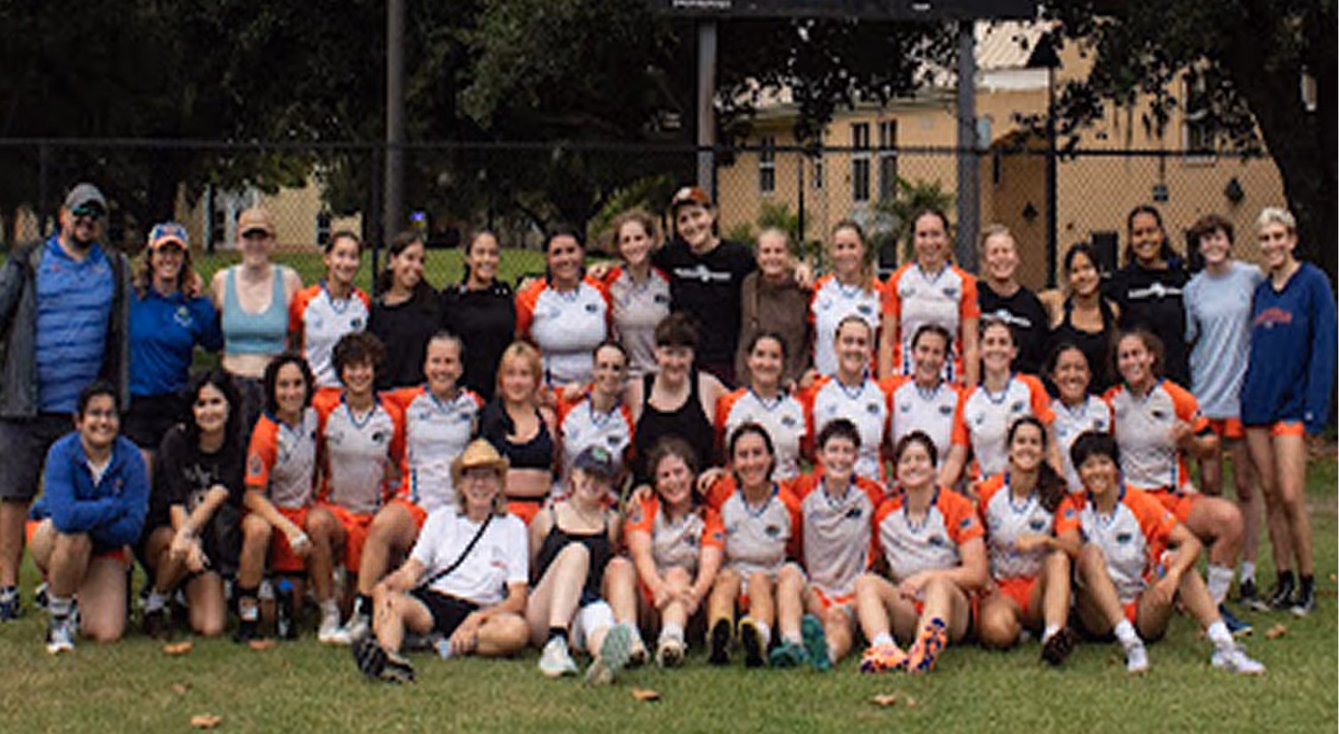Large group of women and a few men posing outdoors on grass in sportswear, some in orange and white uniforms.