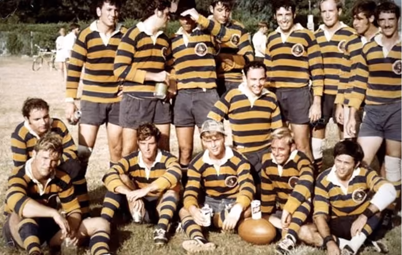Group of rugby players in yellow and black striped jerseys posing with a rugby ball on grass.