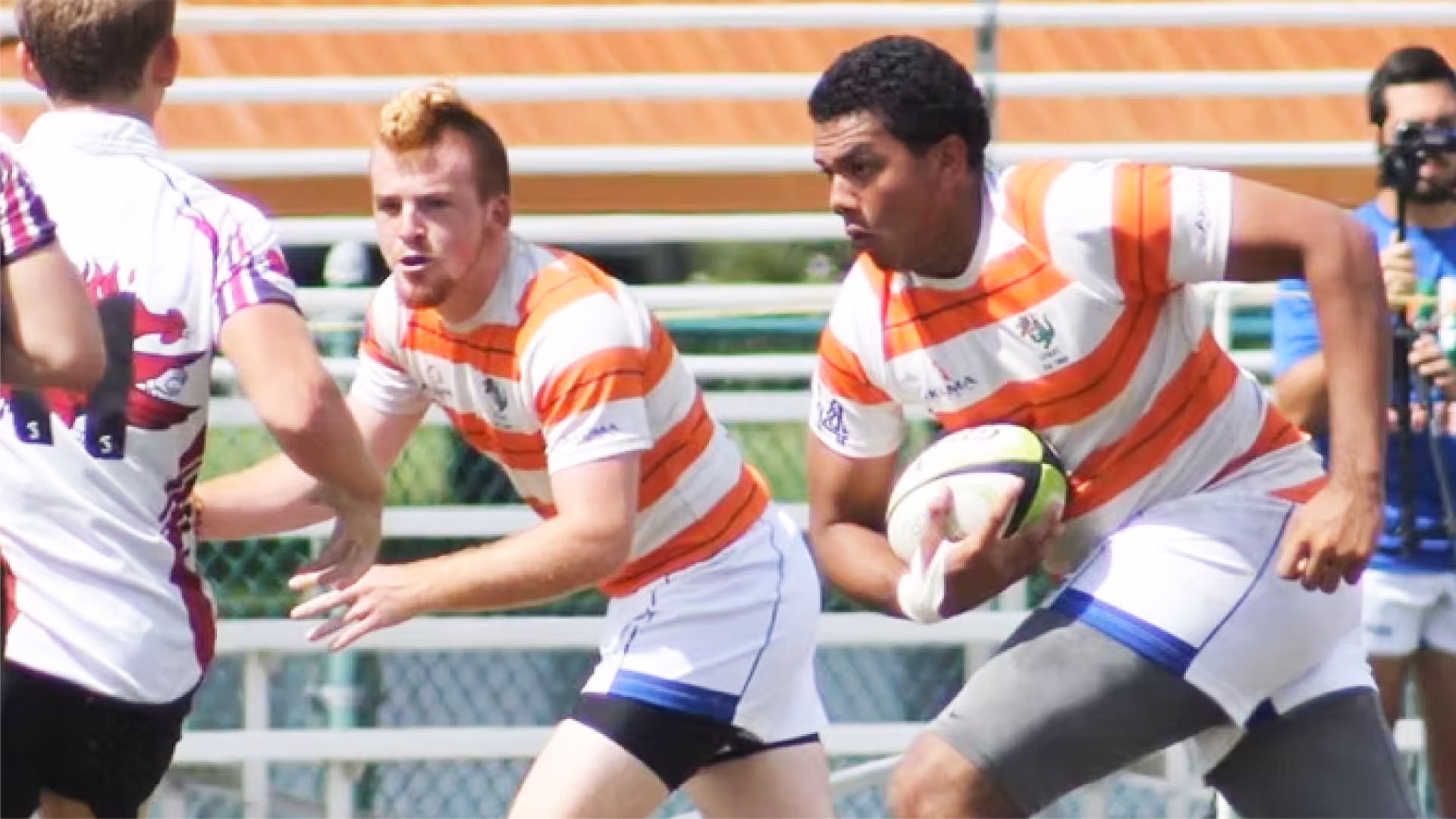 Two rugby players in white and orange striped jerseys running with the ball and preparing to evade a defender in a white jersey with red and purple details during a match.