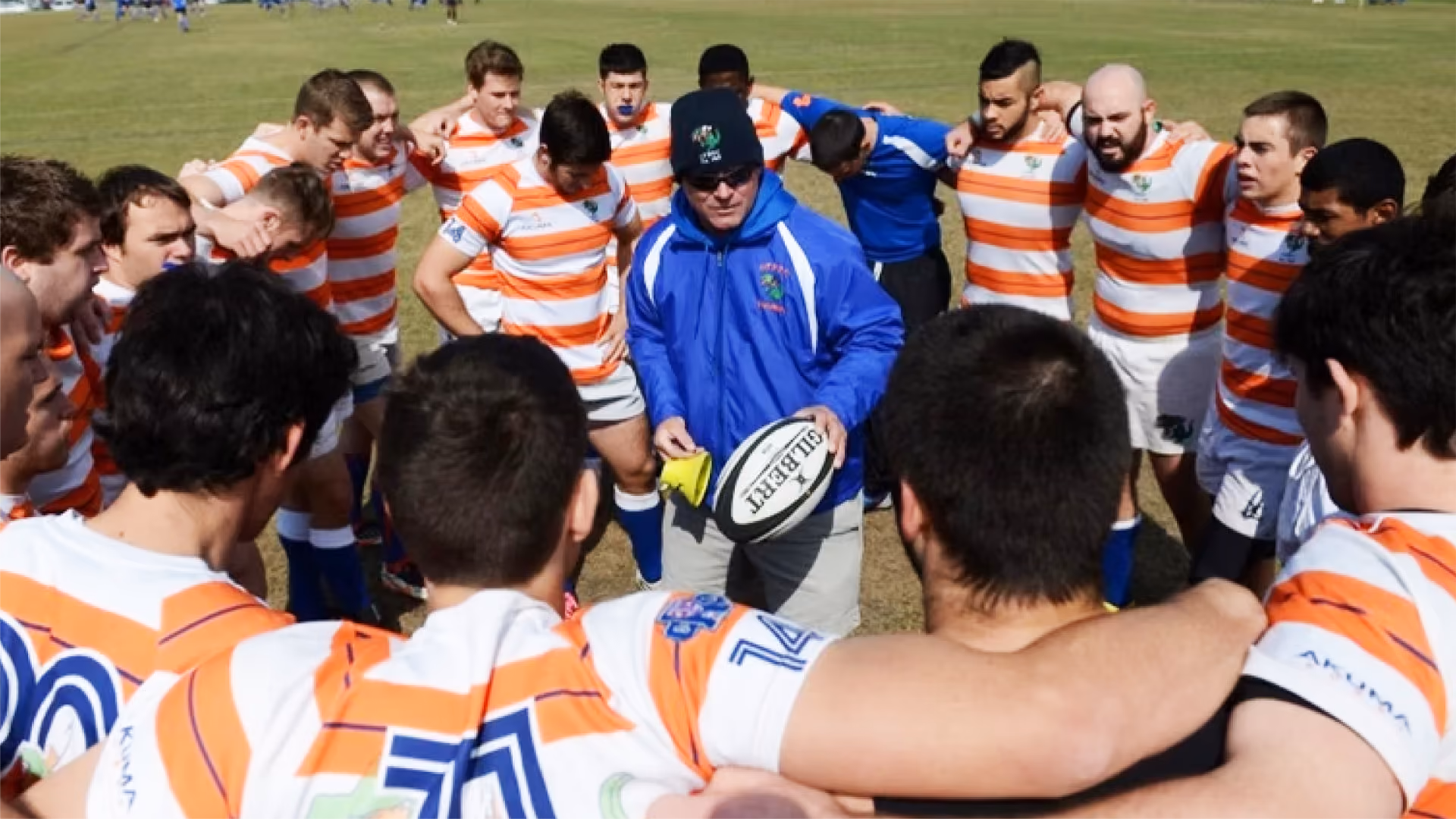 Rugby coach in blue jacket and hat holding a rugby ball talks to his huddled team wearing white jerseys with orange stripes on a grassy field.