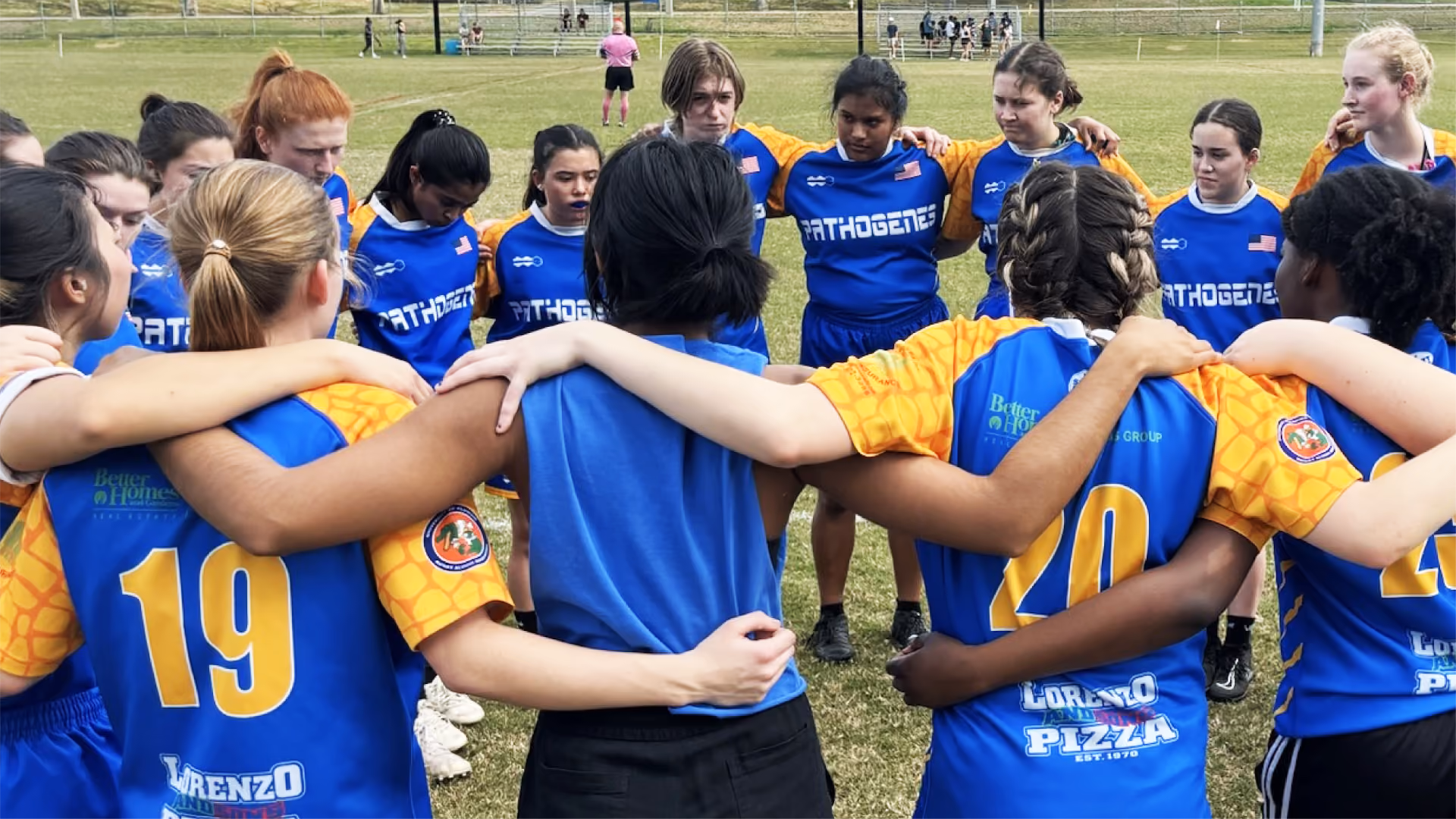 A diverse women's sports team huddled in a circle on a grassy field wearing blue and yellow jerseys with 'PATHOGENS' on the front and numbers on the back.