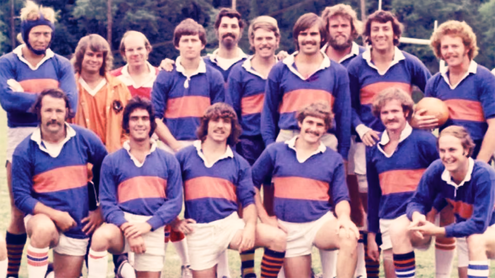 Vintage photo of a rugby team posing outdoors, wearing blue jerseys with an orange stripe and white shorts, some players smiling, one holding a rugby ball.