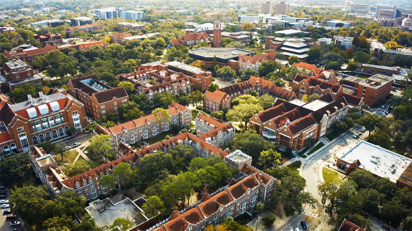 Aerial view of a university campus with red-roofed academic and residential buildings surrounded by trees and roads.