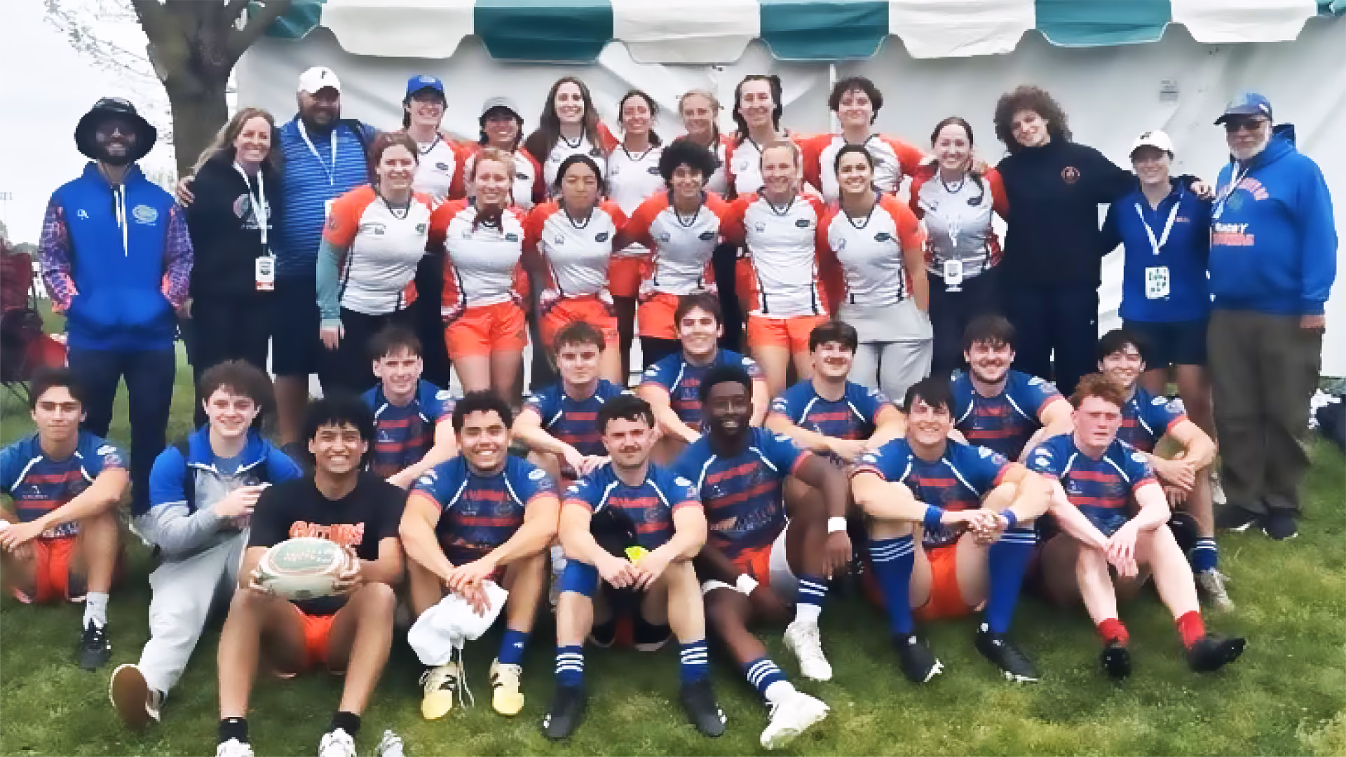 Group photo of a mixed-gender rugby team and their staff, with players wearing blue and orange or white and orange uniforms, gathered outdoors in front of a white tent on green grass.
