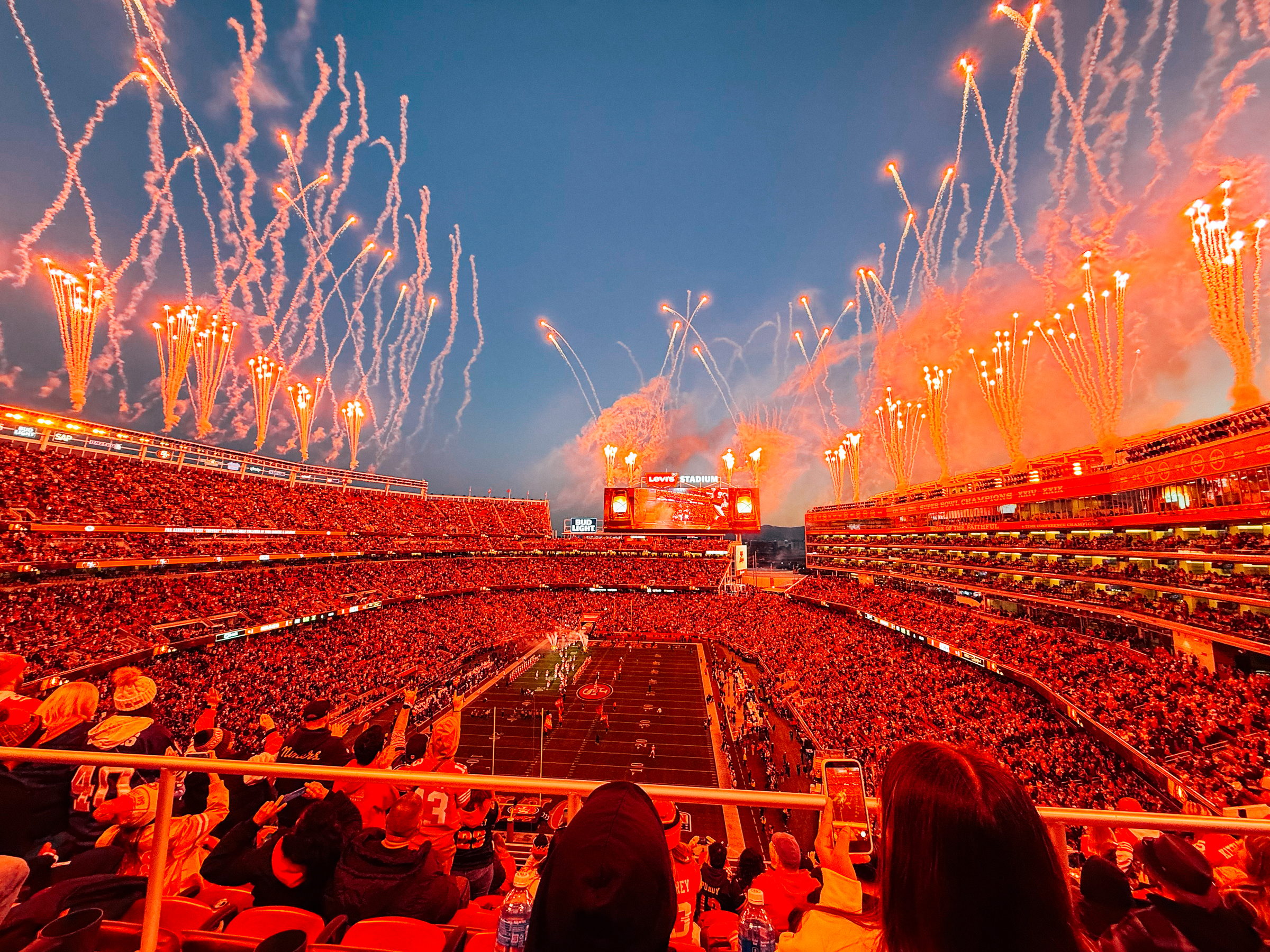 Crowded football stadium with fireworks lighting the sky during a game at sunset.