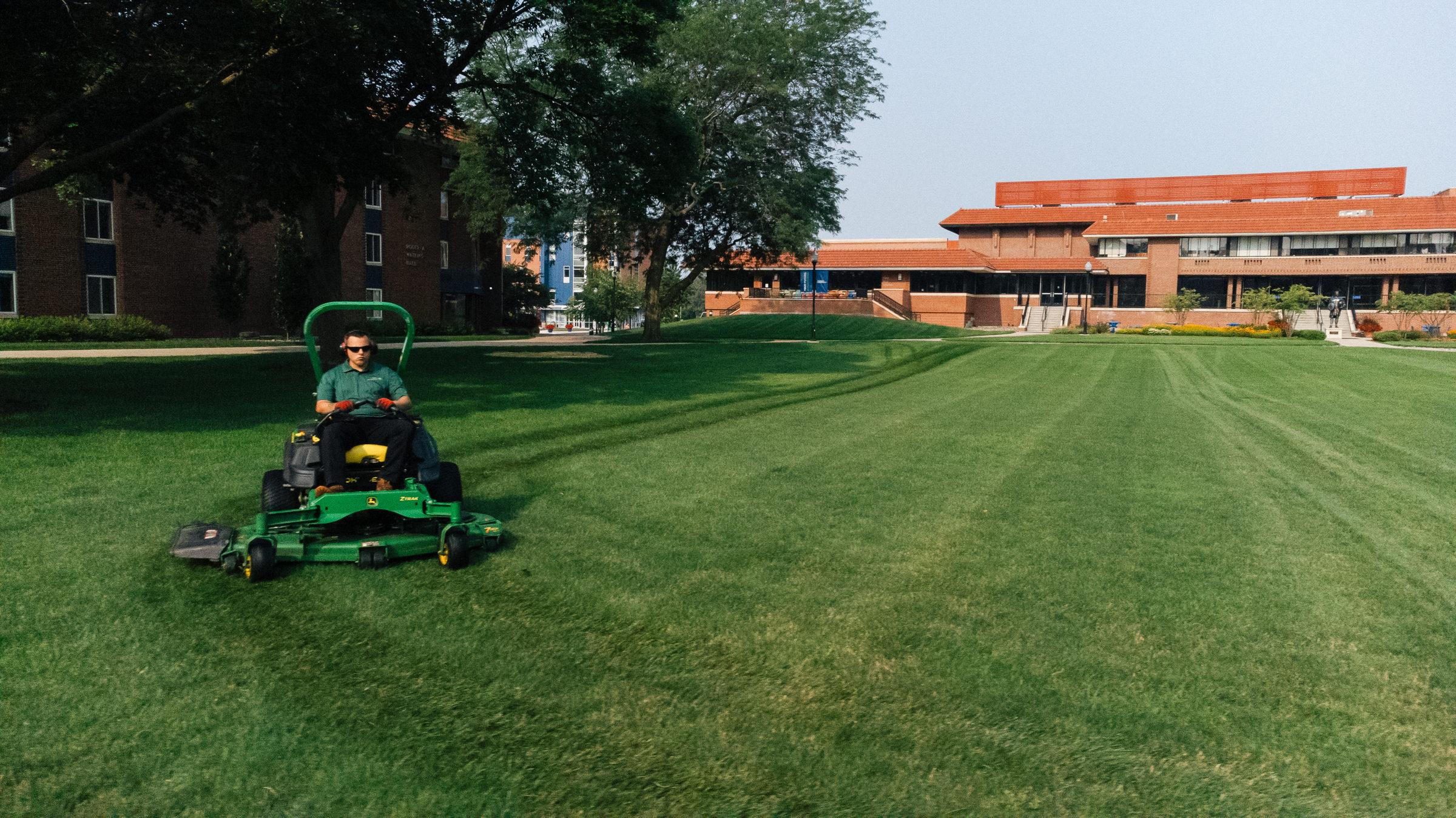 Person riding a green lawn mower cutting grass on a large lawn near brick buildings on a sunny day.