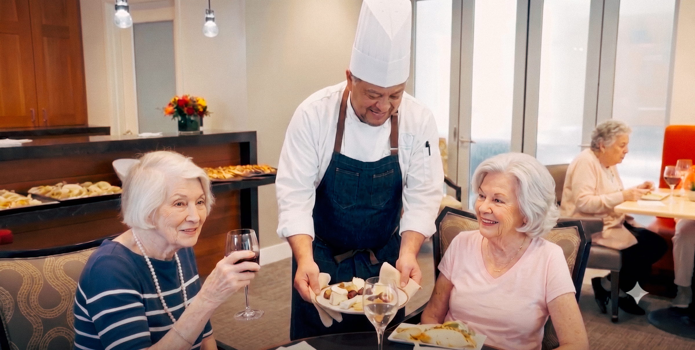Male chef serving food to two elderly women dining and smiling in a cozy restaurant setting.
