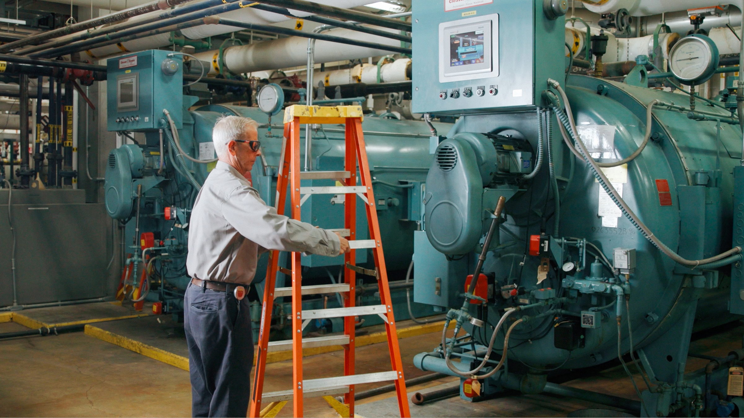 Man in safety glasses setting up orange ladder in industrial machine room with large equipment.