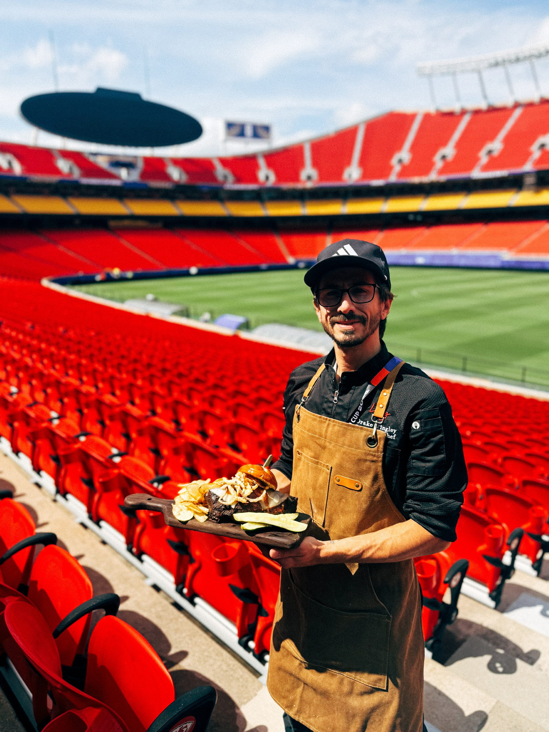 Man wearing apron holding wooden board with sandwich and snacks in a stadium with red seats.