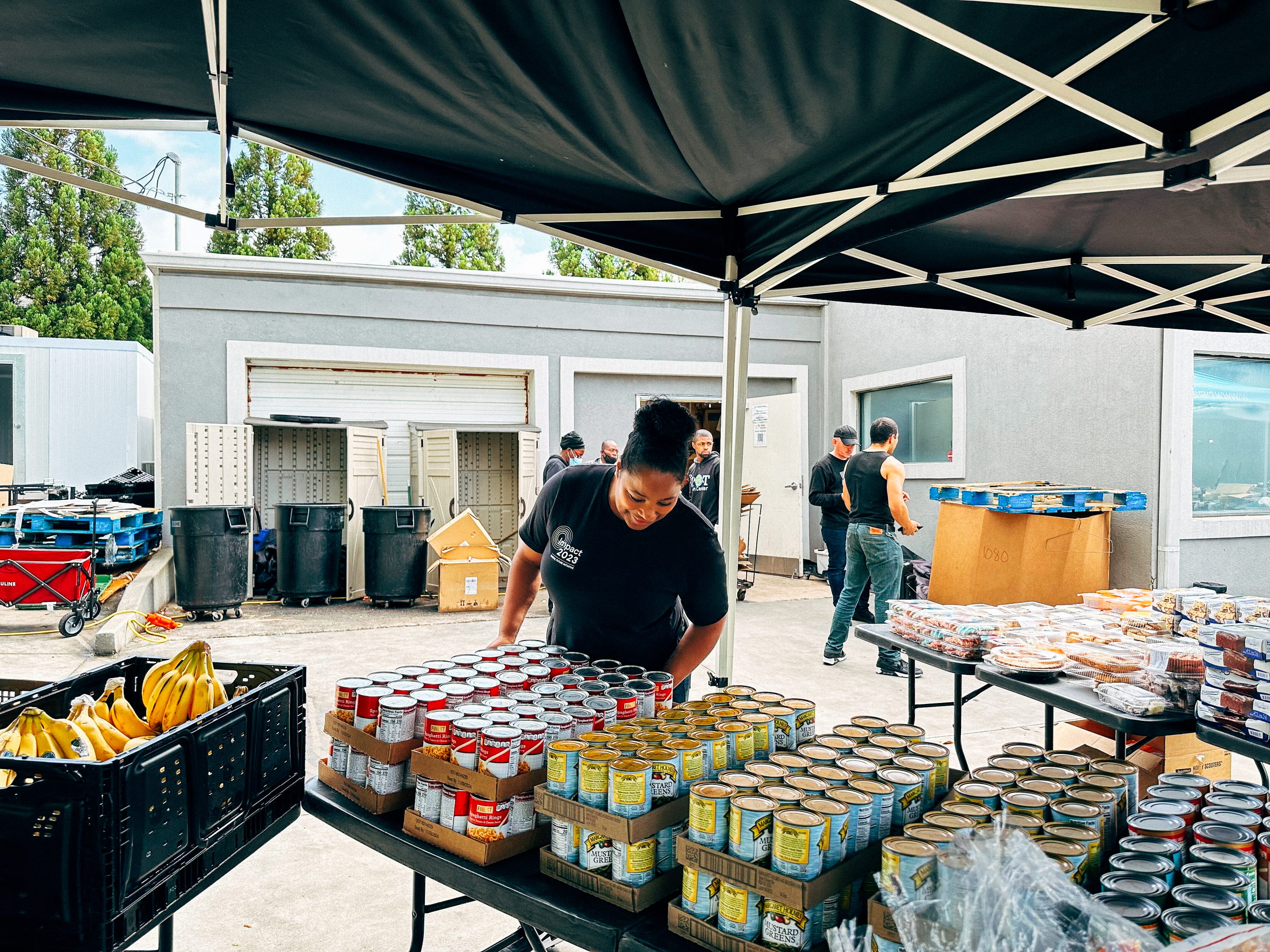 A woman arranges canned goods and bananas under a black canopy at an outdoor food distribution site.