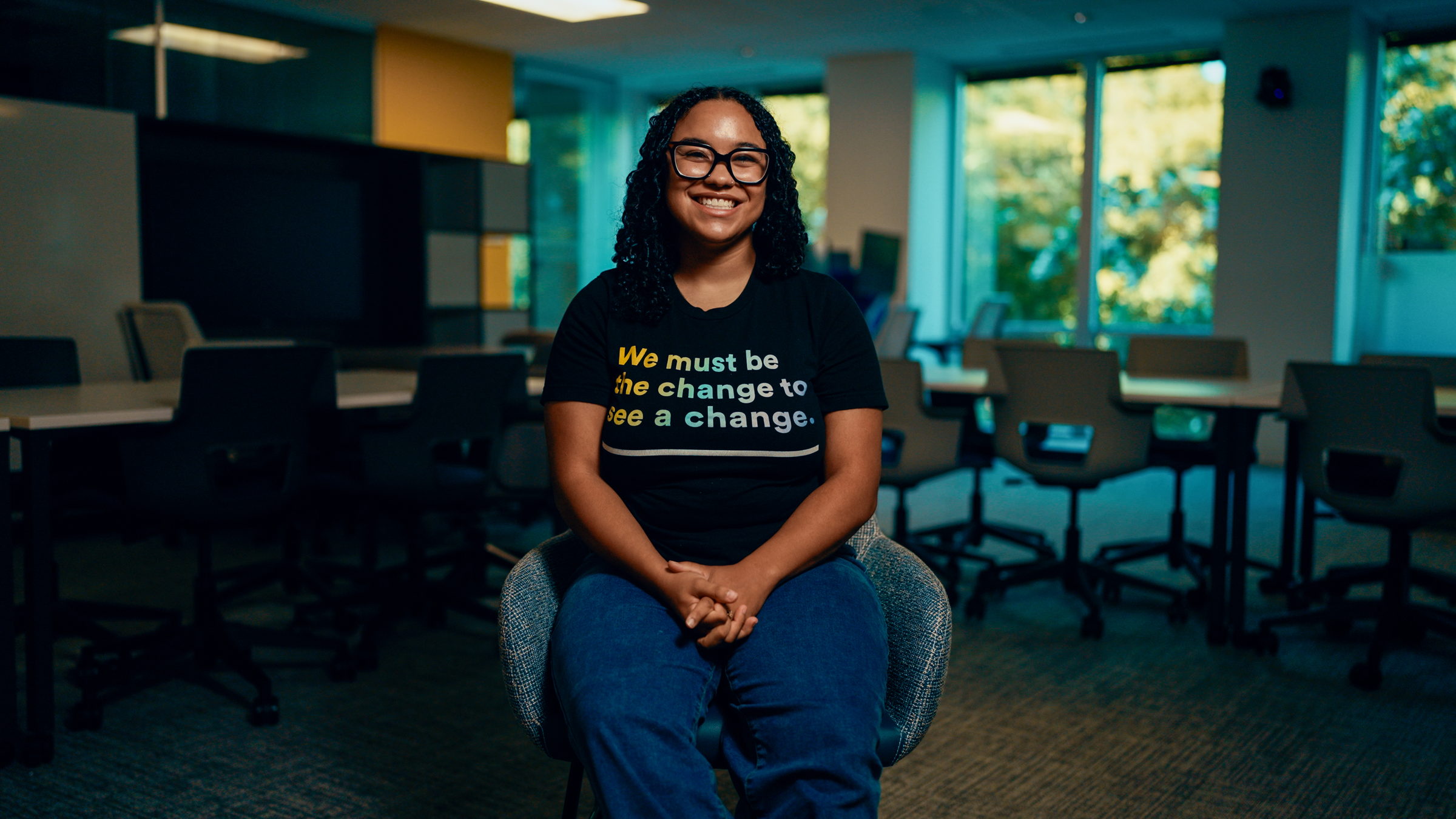 Smiling woman with glasses wearing a black shirt sitting on a chair in a room with tables and chairs.