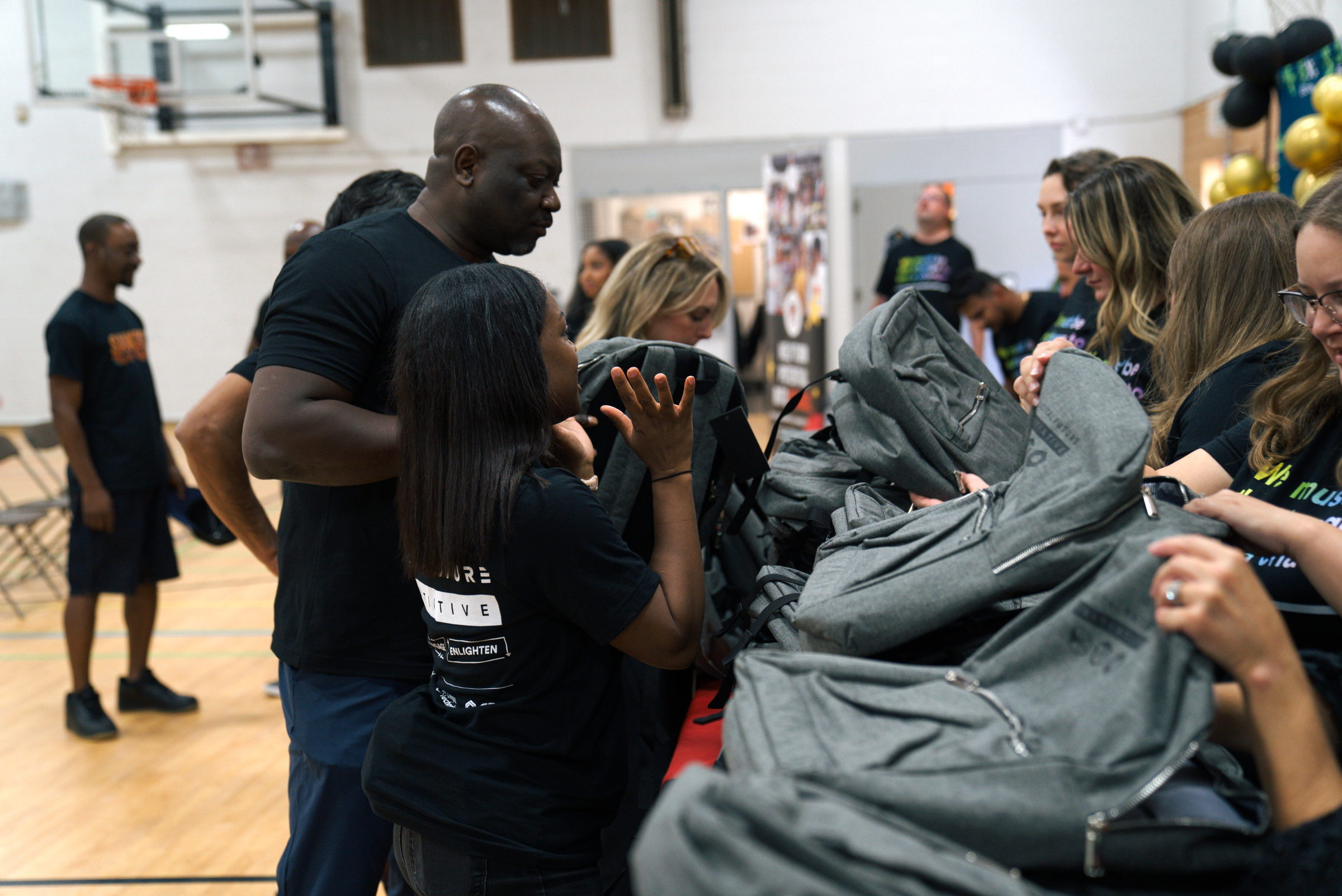 People distributing gray backpacks at a table inside a gym with sports equipment visible.