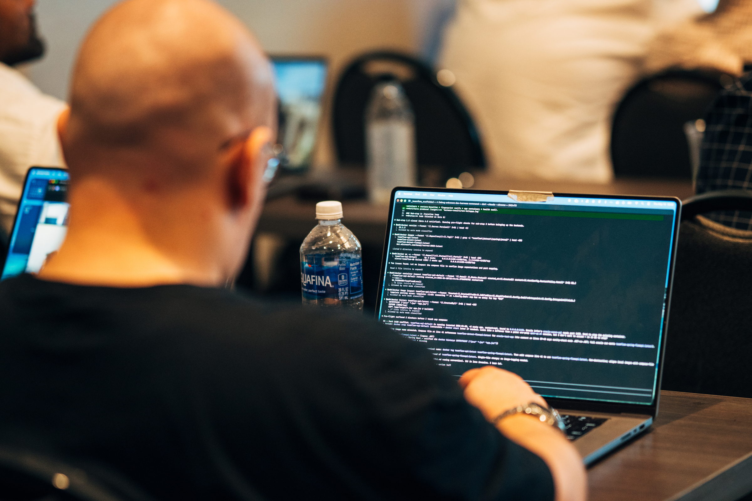 Person with glasses working on a laptop displaying code with a water bottle beside them.