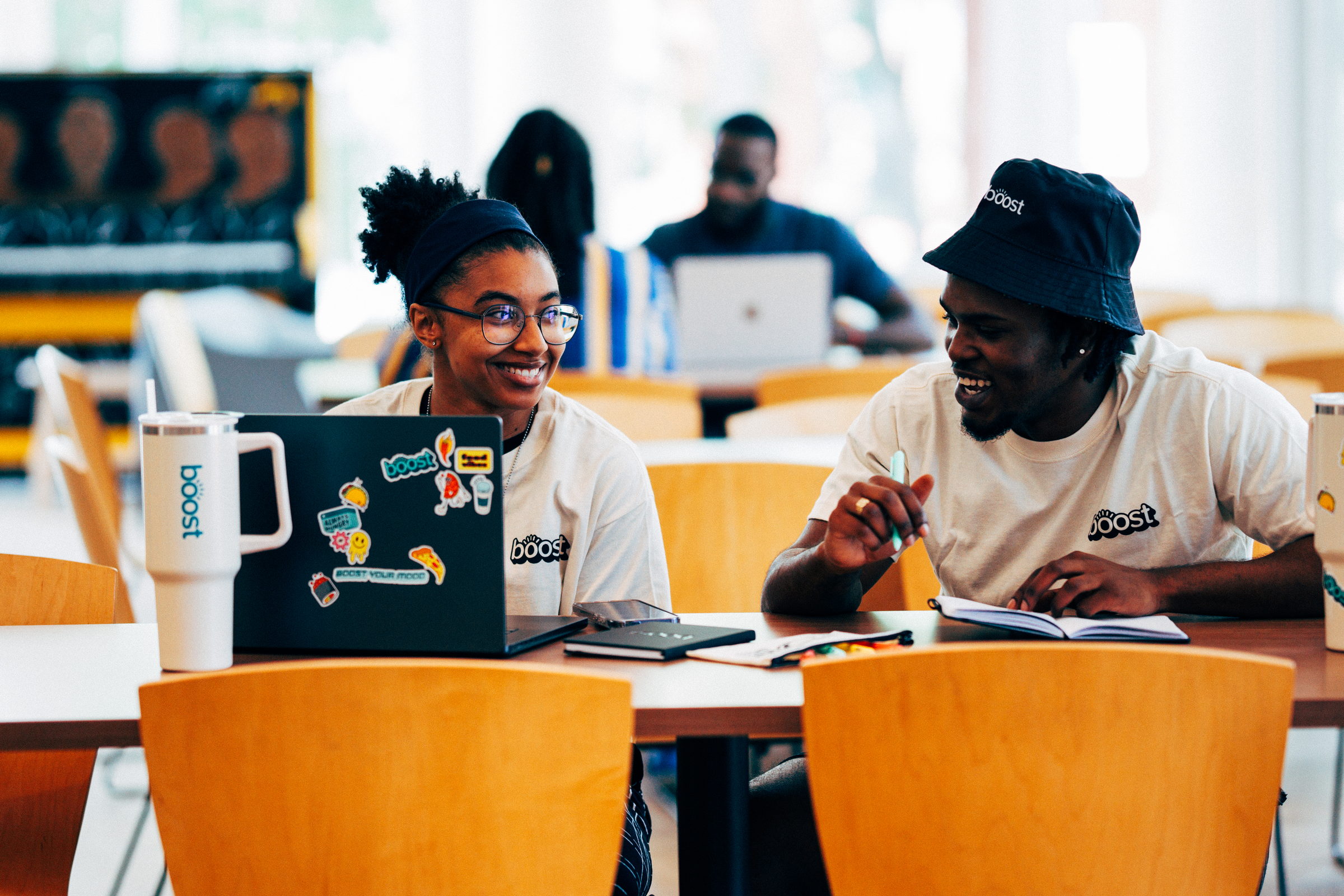 Two people smiling and working together at a table with a laptop and notebooks in a bright room.