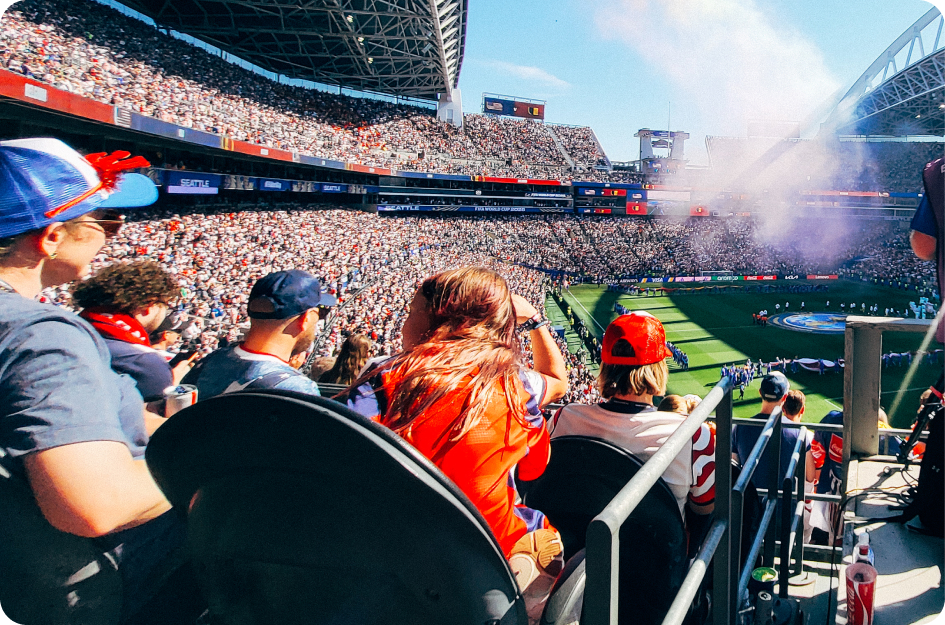 Crowd filling stadium seats at a daytime sporting event with flags, lights, and smoke on the field.