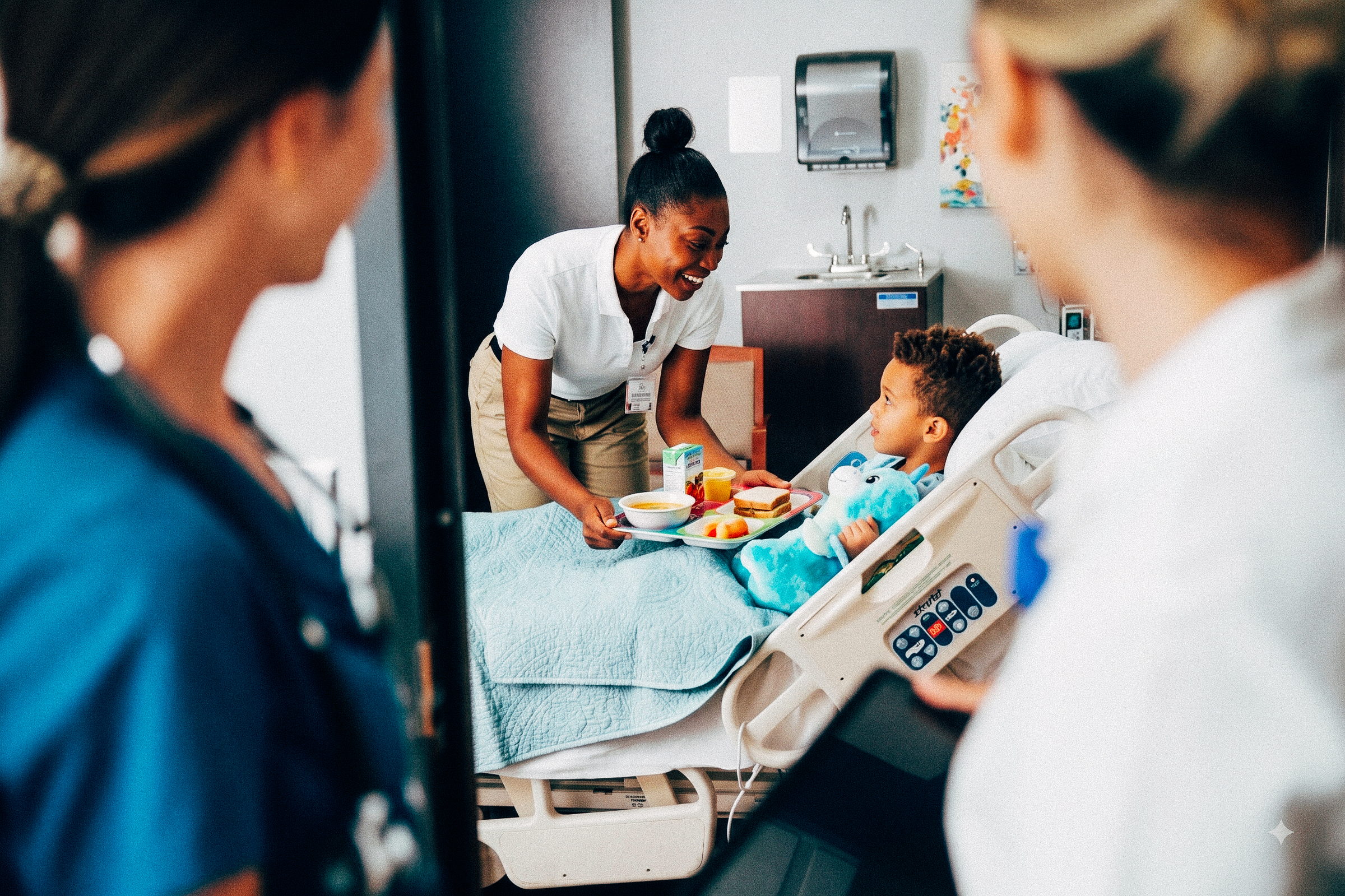 Healthcare worker in white shirt handing meal tray to a child patient holding a blue stuffed animal in hospital bed.