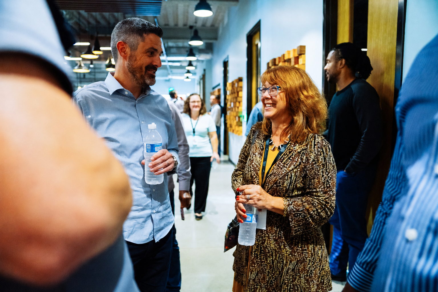 Man and woman holding water bottles and talking in a busy indoor hallway with people in the background.