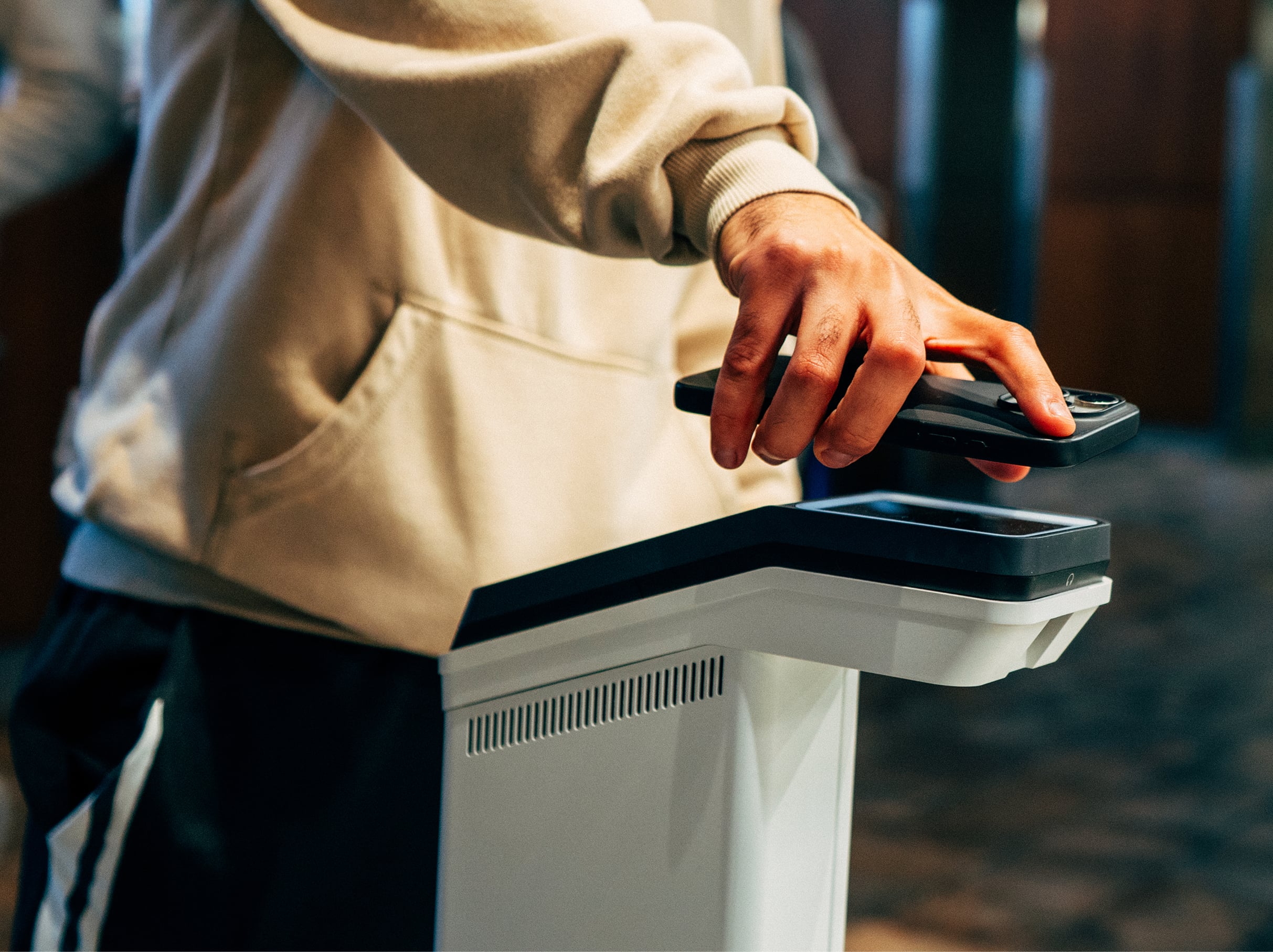Person in beige hoodie holding phone over a white digital kiosk.