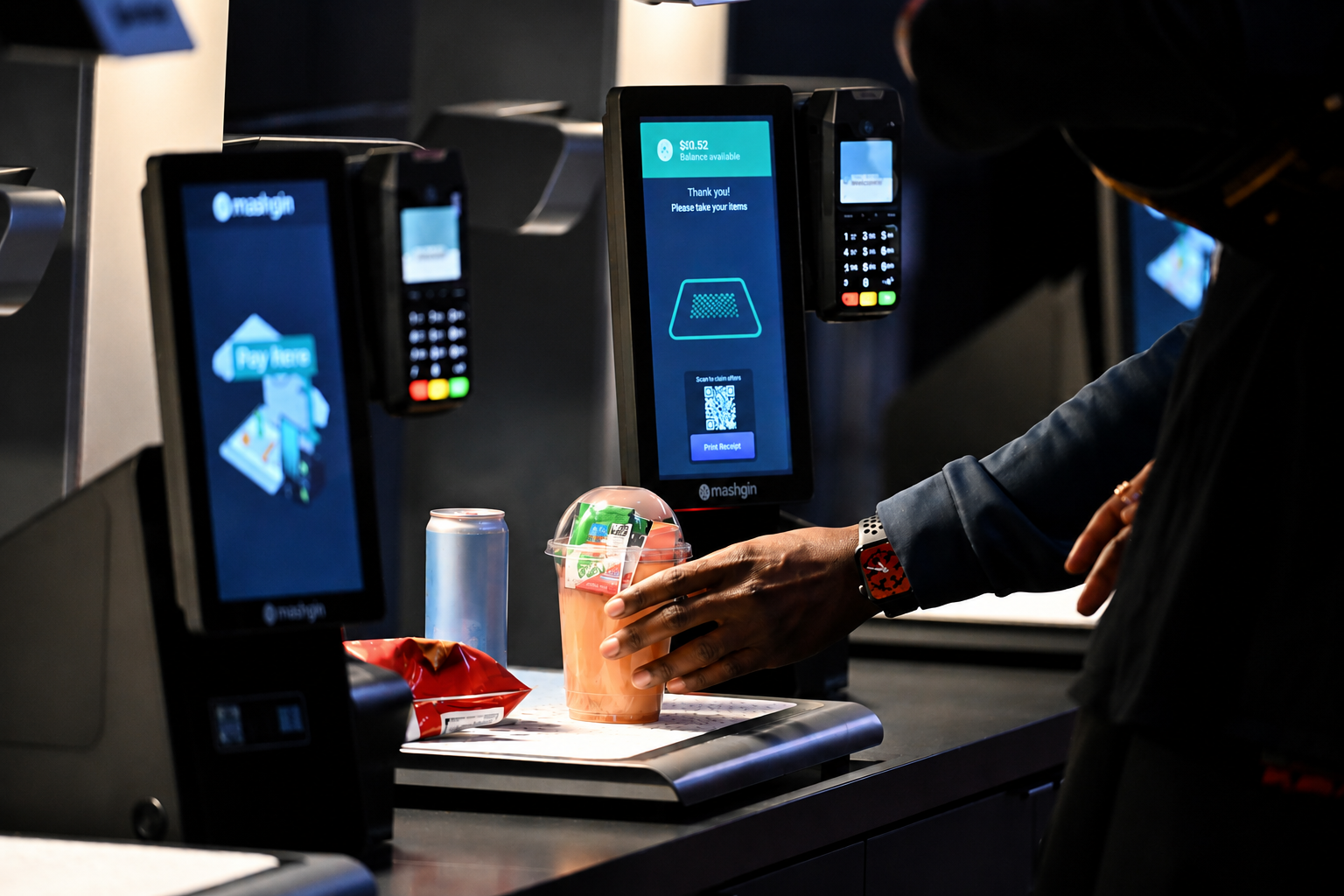 Hand placing a drink, chip bag, and can on a self-checkout counter with a digital screen.