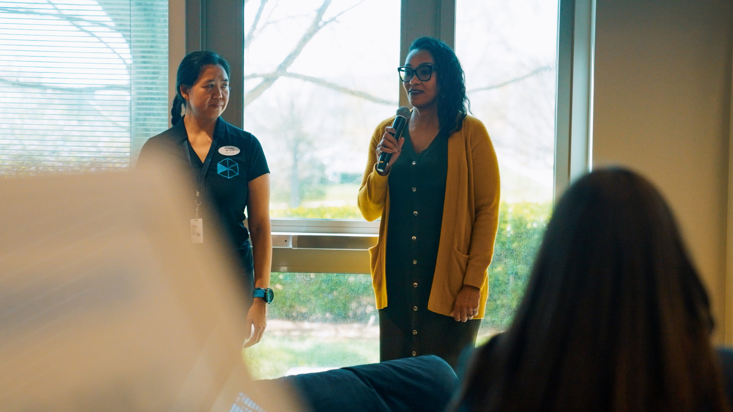 Two women standing by a window; one speaking into a microphone, the other listening attentively.