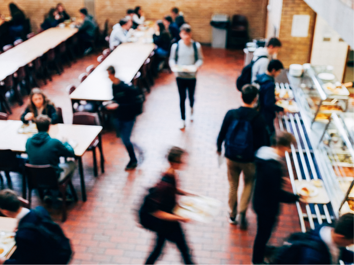 Blurred cafeteria scene with students carrying trays and others seated at tables eating.