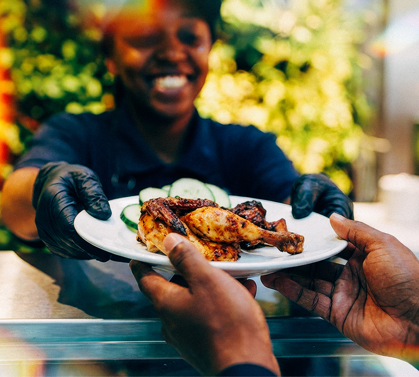 Server wearing gloves hands a plate with grilled chicken and cucumber slices to a customer.