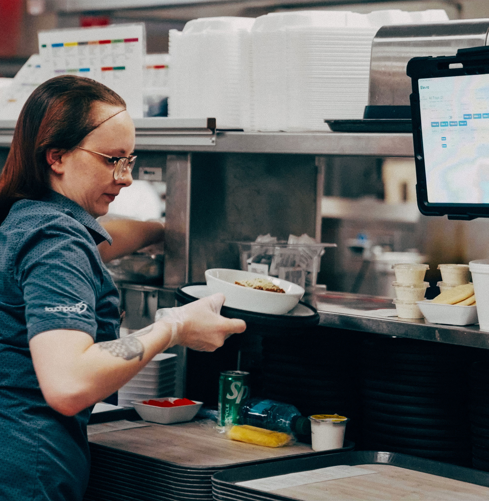 Woman in a blue shirt preparing food on a kitchen pass with trays and a mounted tablet.