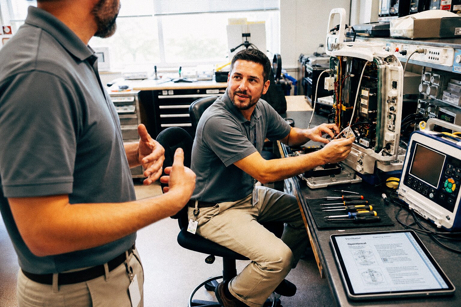 Two technicians collaborate over an open machine, using tools and a tablet with an open manual.