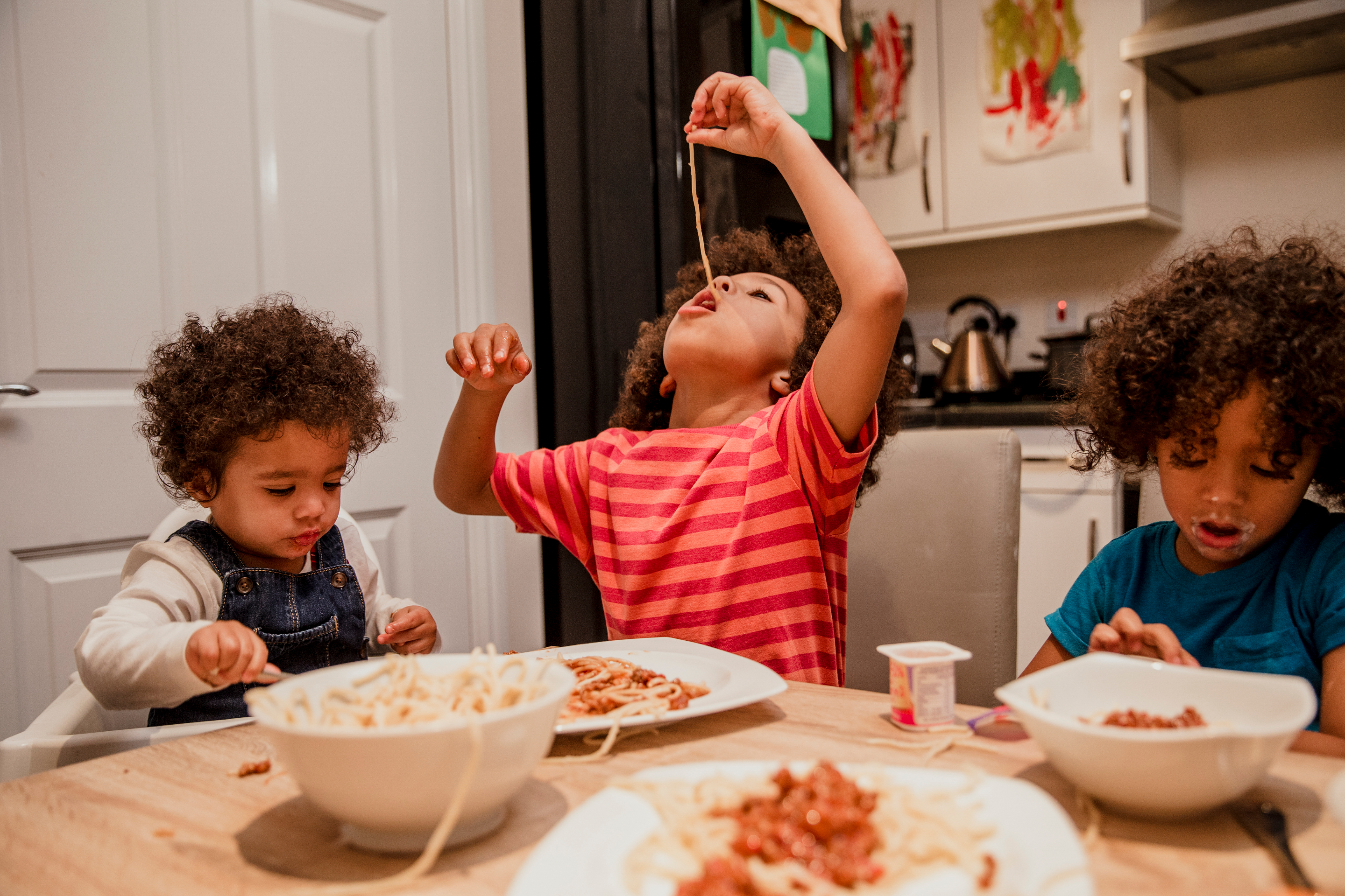 Three children eating spaghetti, with the middle child lifting a noodle